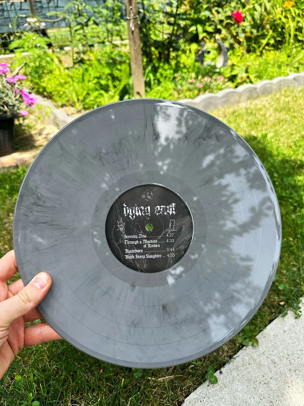 Person holding a gray vinyl record with a black label, outdoors in a garden with flowers and greenery in the background.