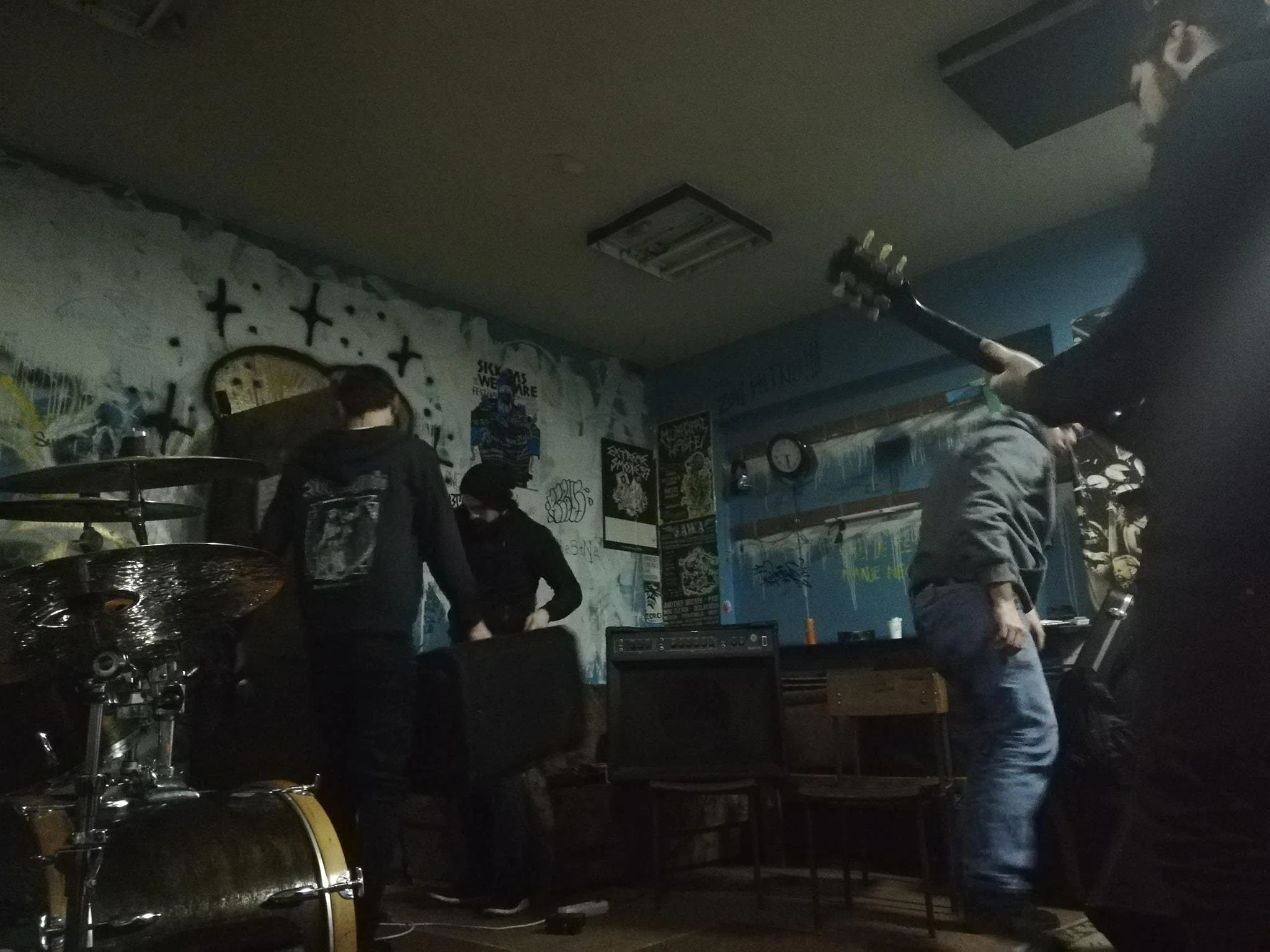 A band playing music in a dimly lit room with graffiti on the wall, with a person holding a guitar in the foreground.