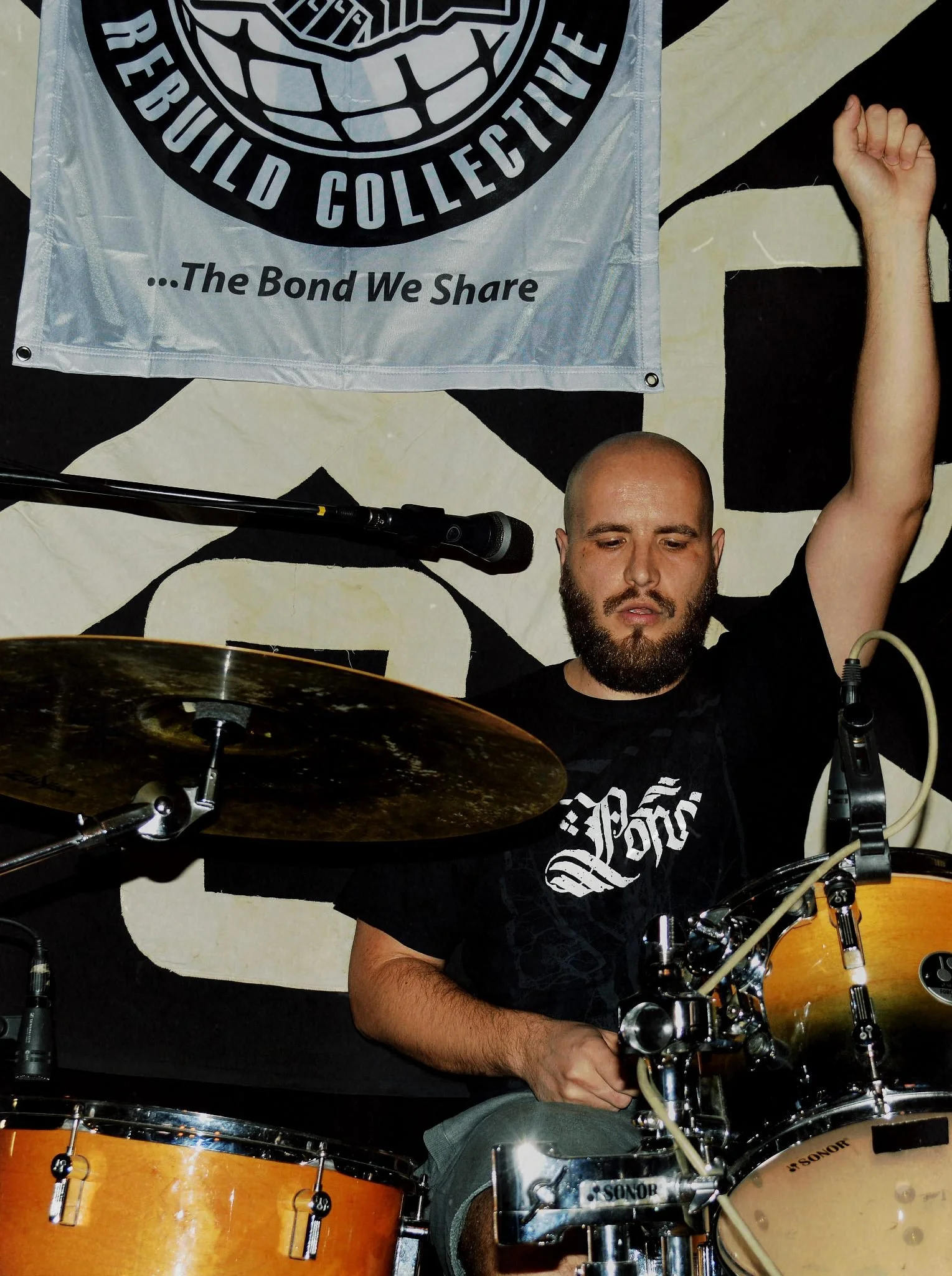 A man with a beard and bald head playing a drum set, with a banner in the background. The man is wearing a black T-shirt with white text and has his arm raised while playing. The drum set includes a cymbal and a bass drum.
