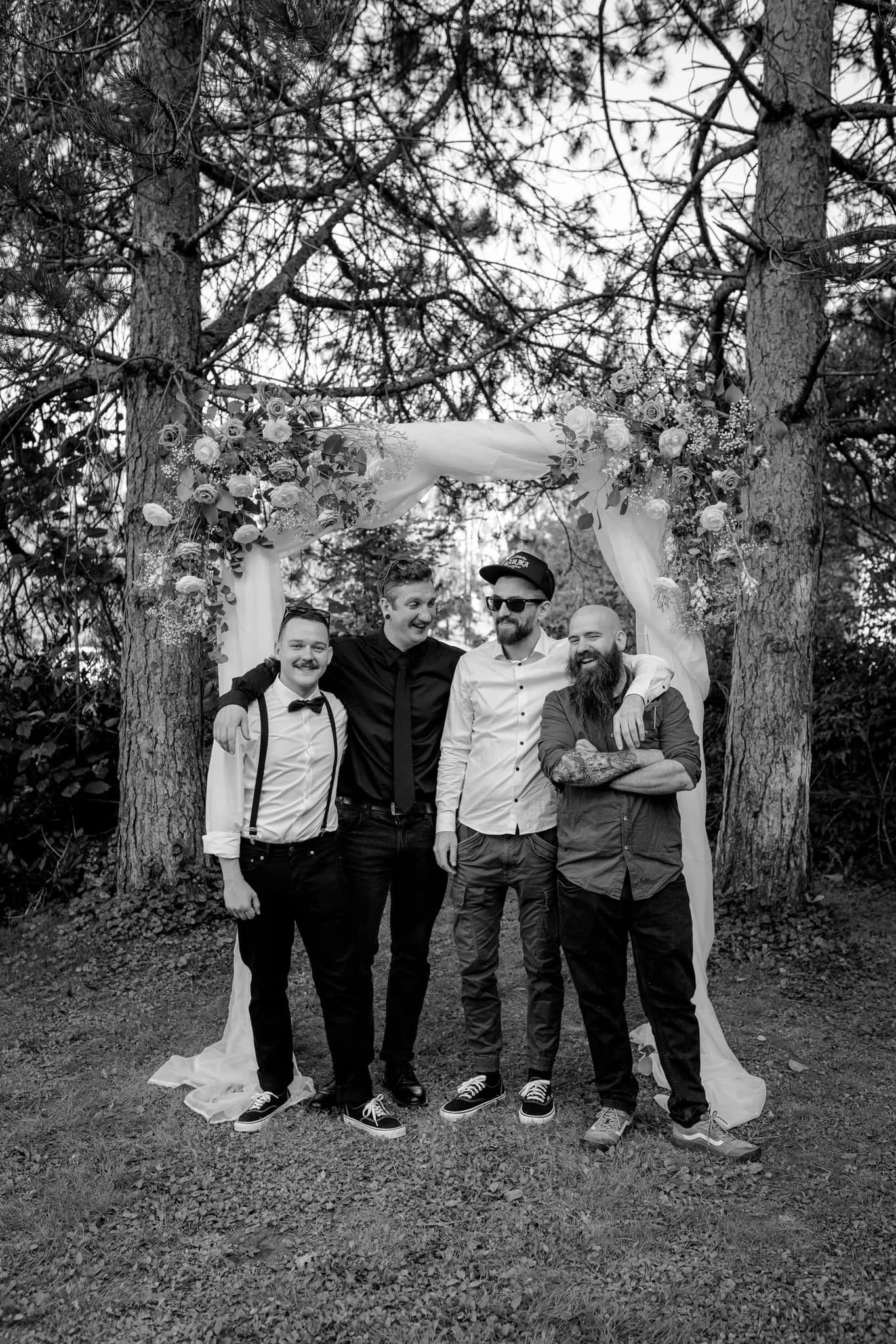A black and white photo of four men standing outdoors in front of trees with a floral wedding arch. The men are smiling and have their arms around each other, dressed in semi-formal attire, with some wearing suspenders, a bow tie, sunglasses, and a h