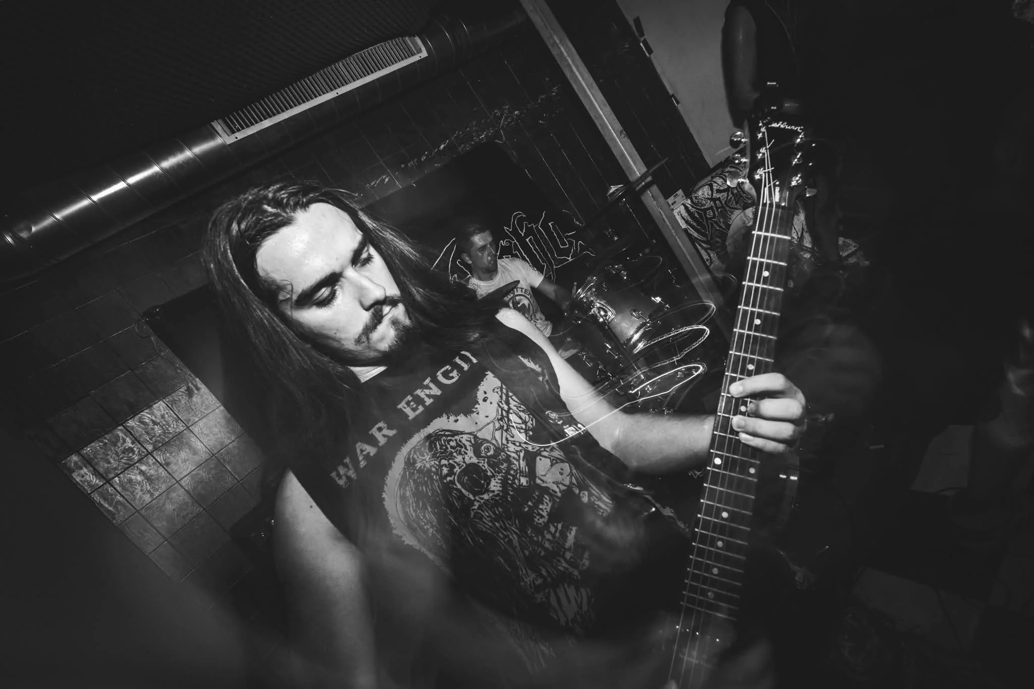 A black-and-white photo of a young man with long hair playing an electric guitar, with another person in the background, in a bar or music venue setting.