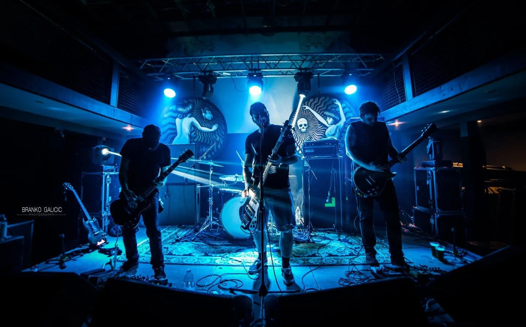 A band performing live on stage under blue lights, with four musicians playing guitars and drums, and a colorful artwork backdrop.