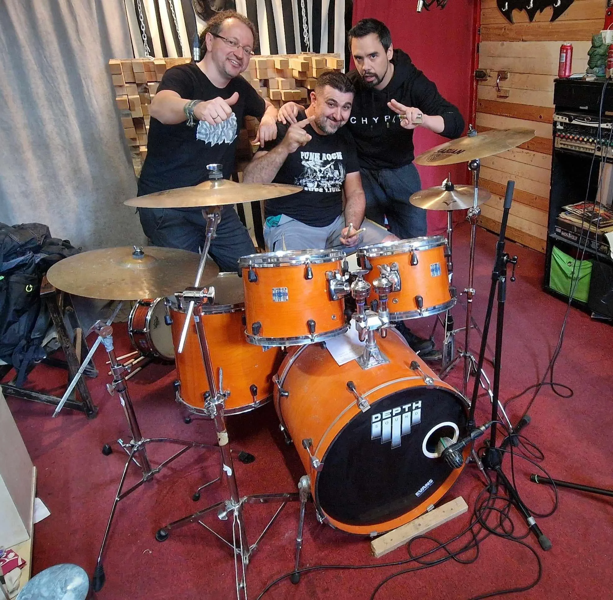 Three men in a room with a drum set, recording equipment, and guitars hanging on the wall, posing and pointing at the camera. One man is sitting at the drum kit, and the other two are standing behind him, making gestures.