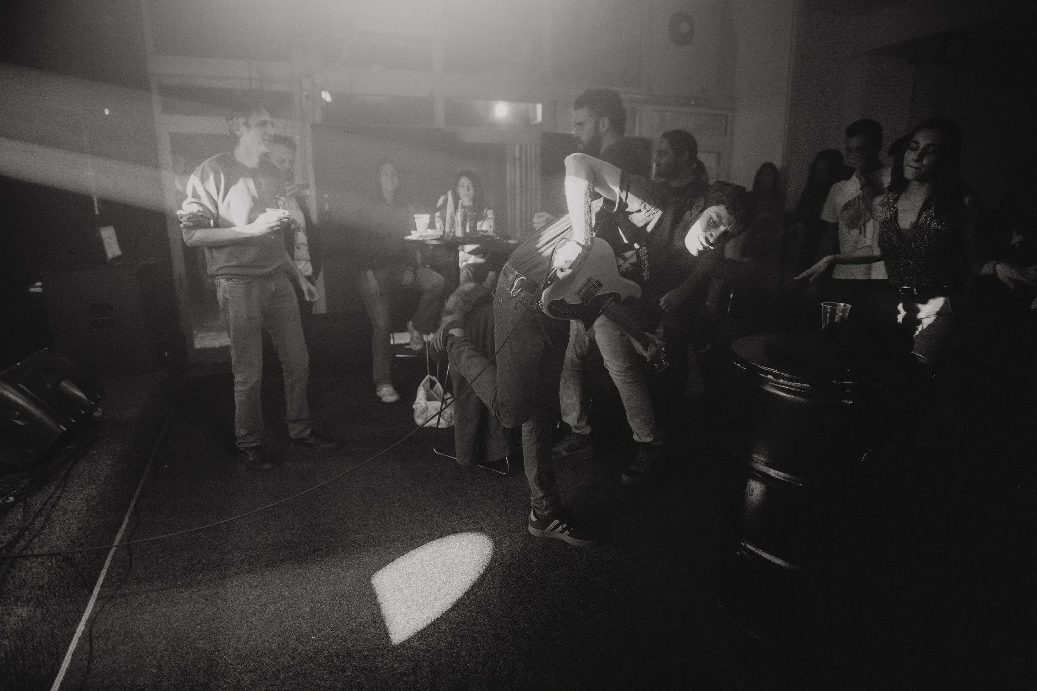 A group of people gathered in a dimly lit room, with some standing and others sitting, as a person with a guitar performs. The scene has a casual, social atmosphere.