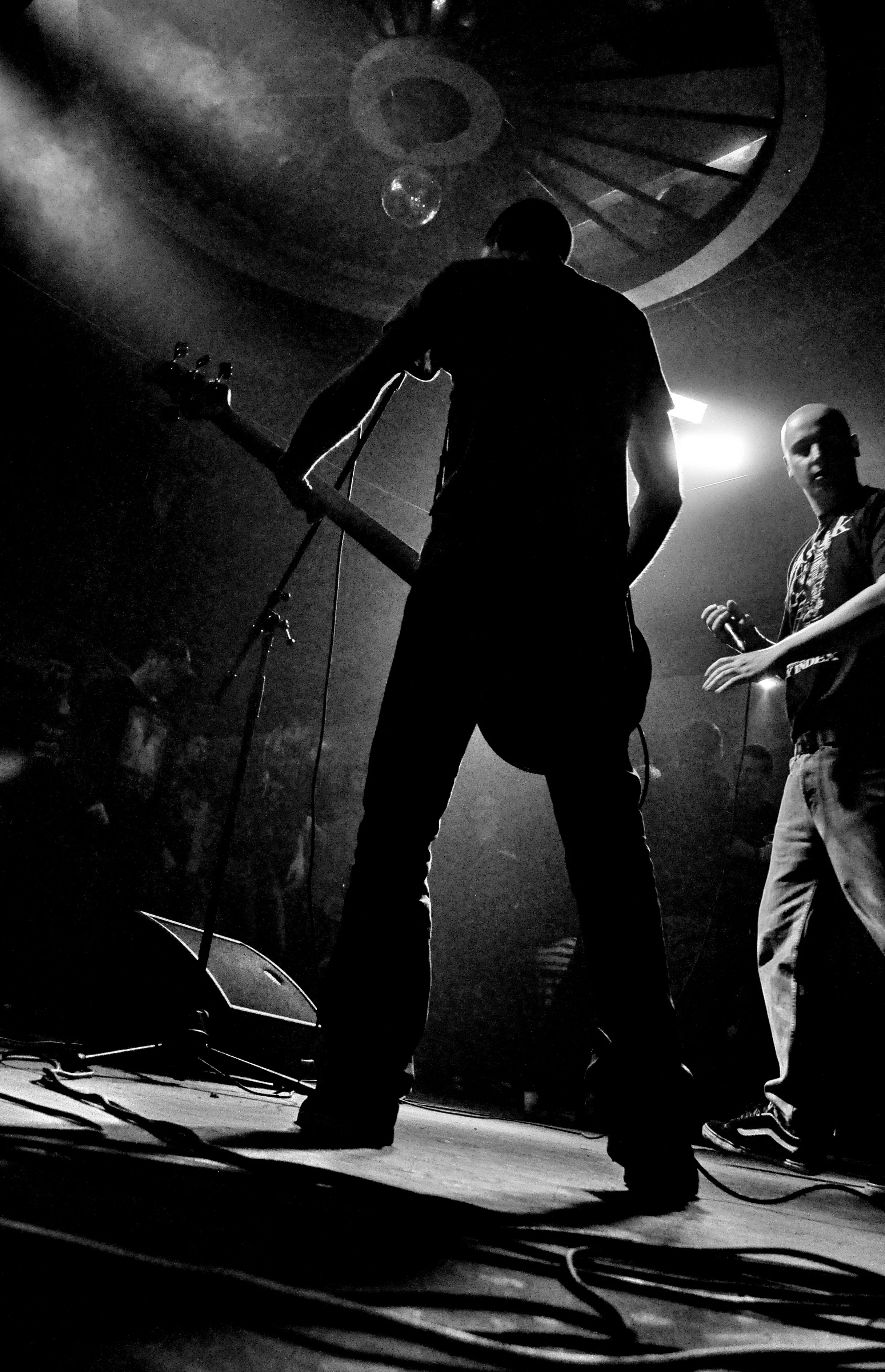 Silhouettes of two musicians performing on stage with microphones and musical instruments, backlit by stage lights, in a smoky live music venue.