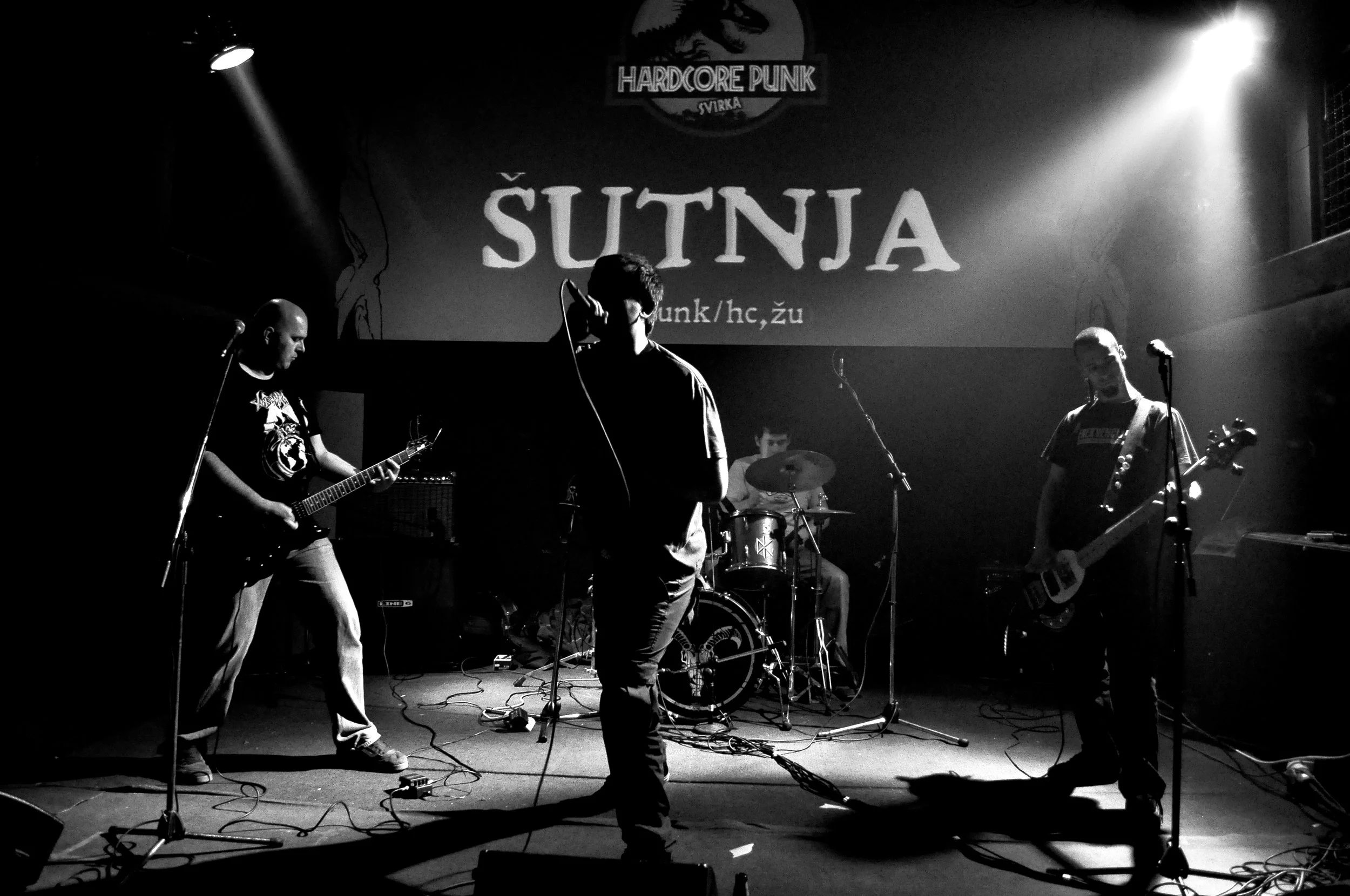A live music band performing on stage with four members, including a guitarist, a bassist, a drummer, and a vocalist, in a dark setting with spotlights, with a large screen behind them displaying the words 'ŠUTNJA' and 'HARDCORE PUNK'.