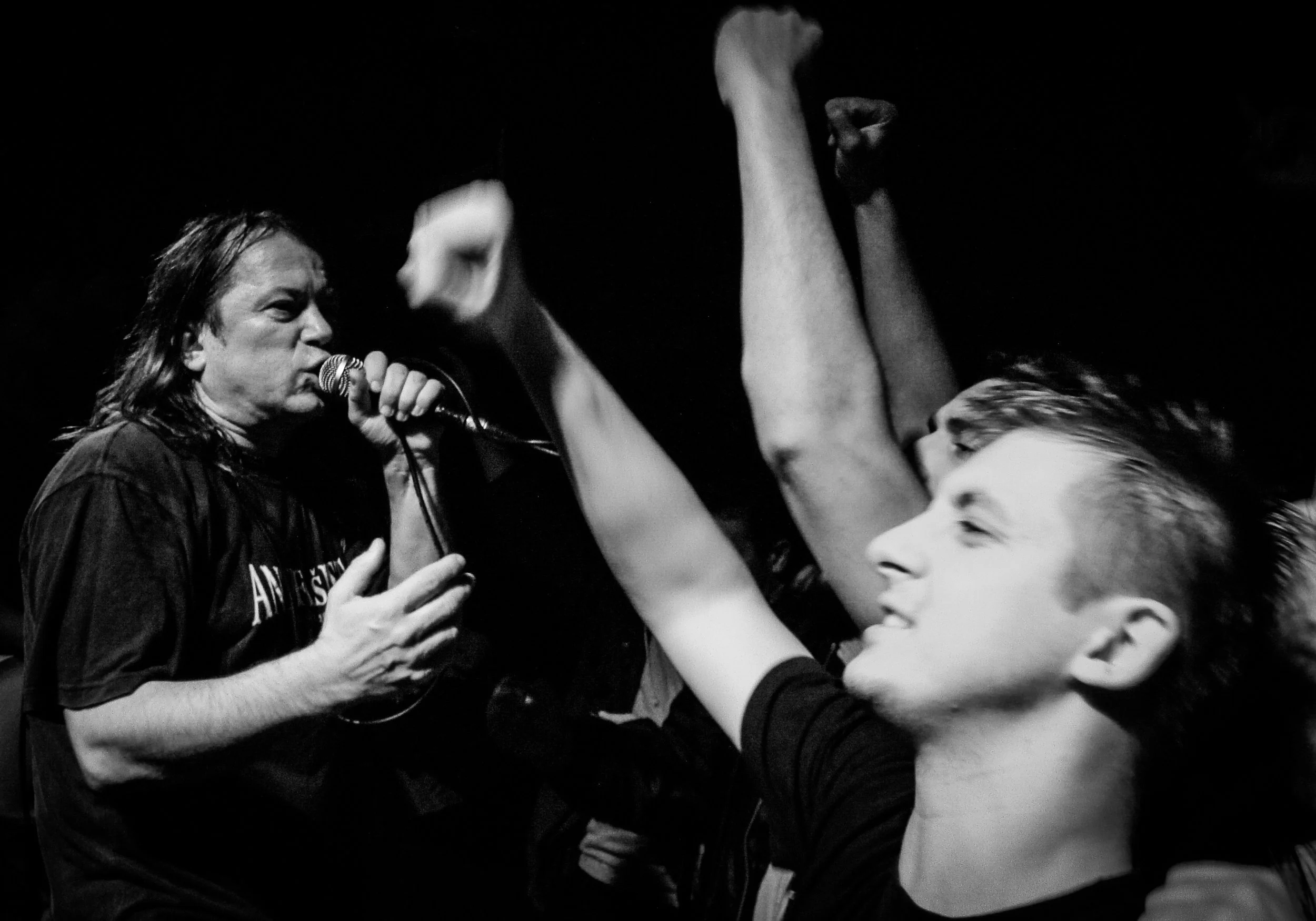 A woman singing into a microphone while engaging with a young audience member with outstretched arms, in a dark setting. Band name: Kud Idijoti