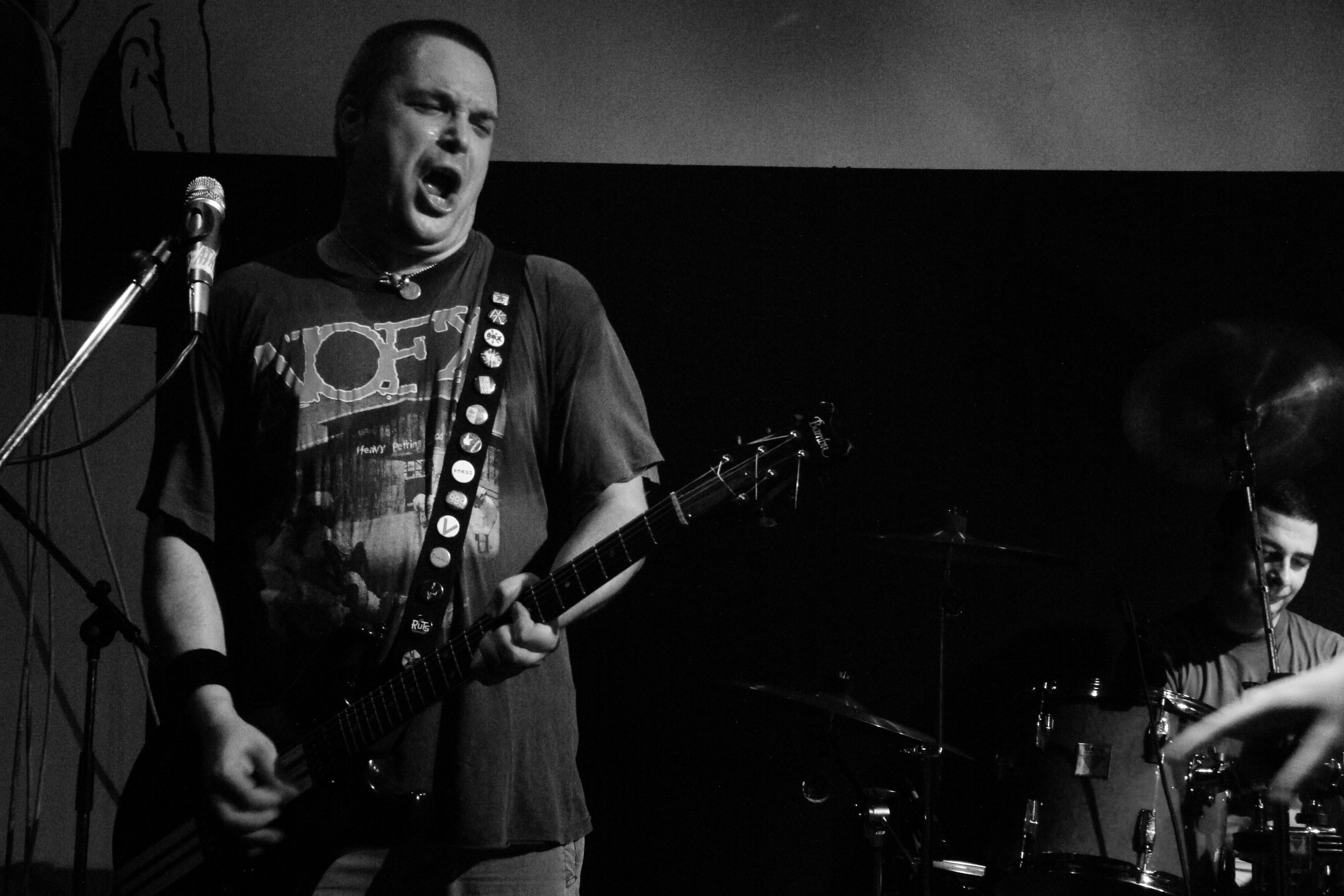 A black and white photo of a male musician passionately singing into a microphone while playing an electric guitar on a stage. There are drums and a female musician in the background. Band name: Atheist Rap