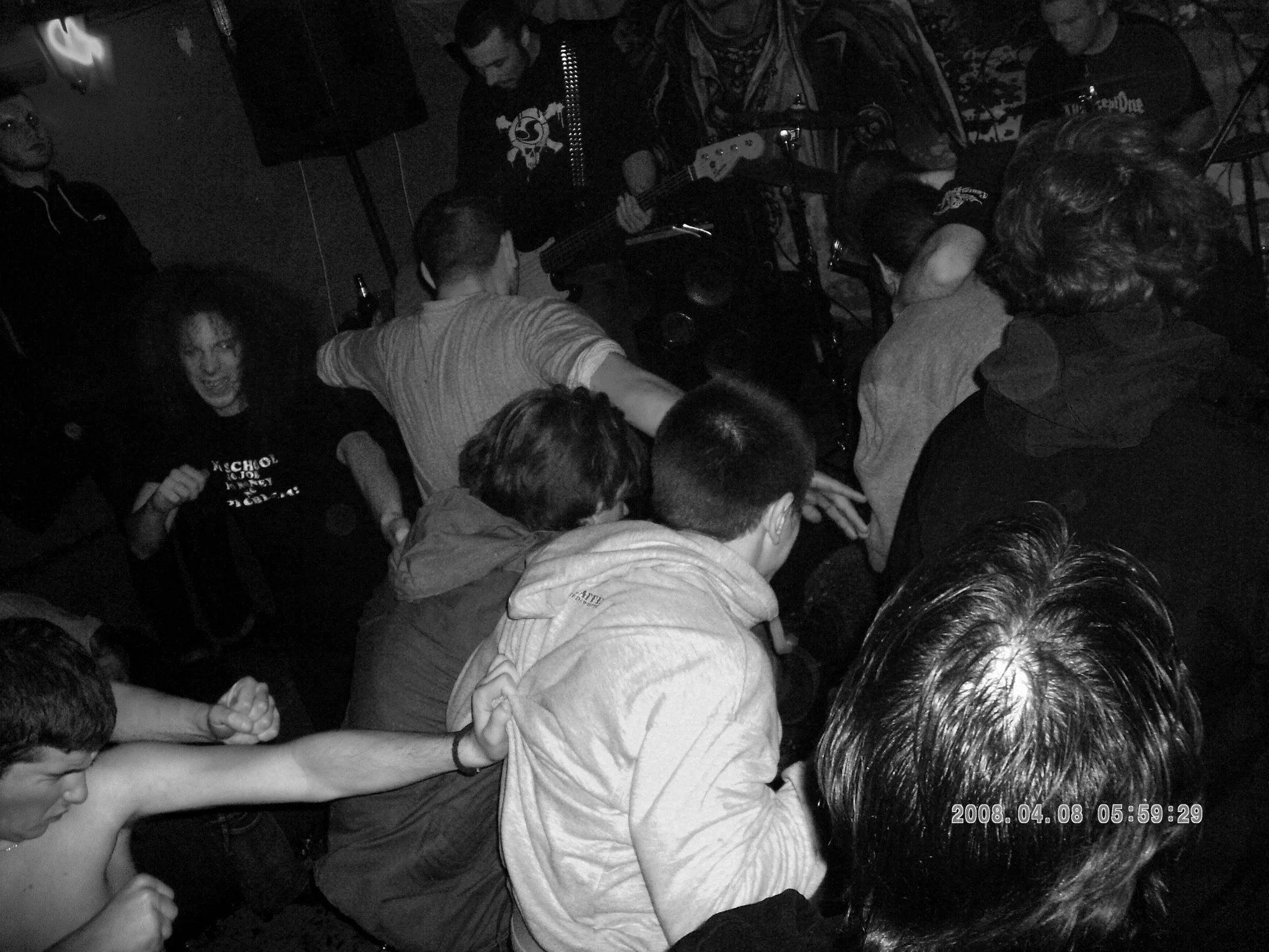A lively indoor scene of a music gig with multiple people tightly packed together, some reaching out or touching others, watching a band perform on stage. The image is in black and white, showing the crowd's energy and engagement.