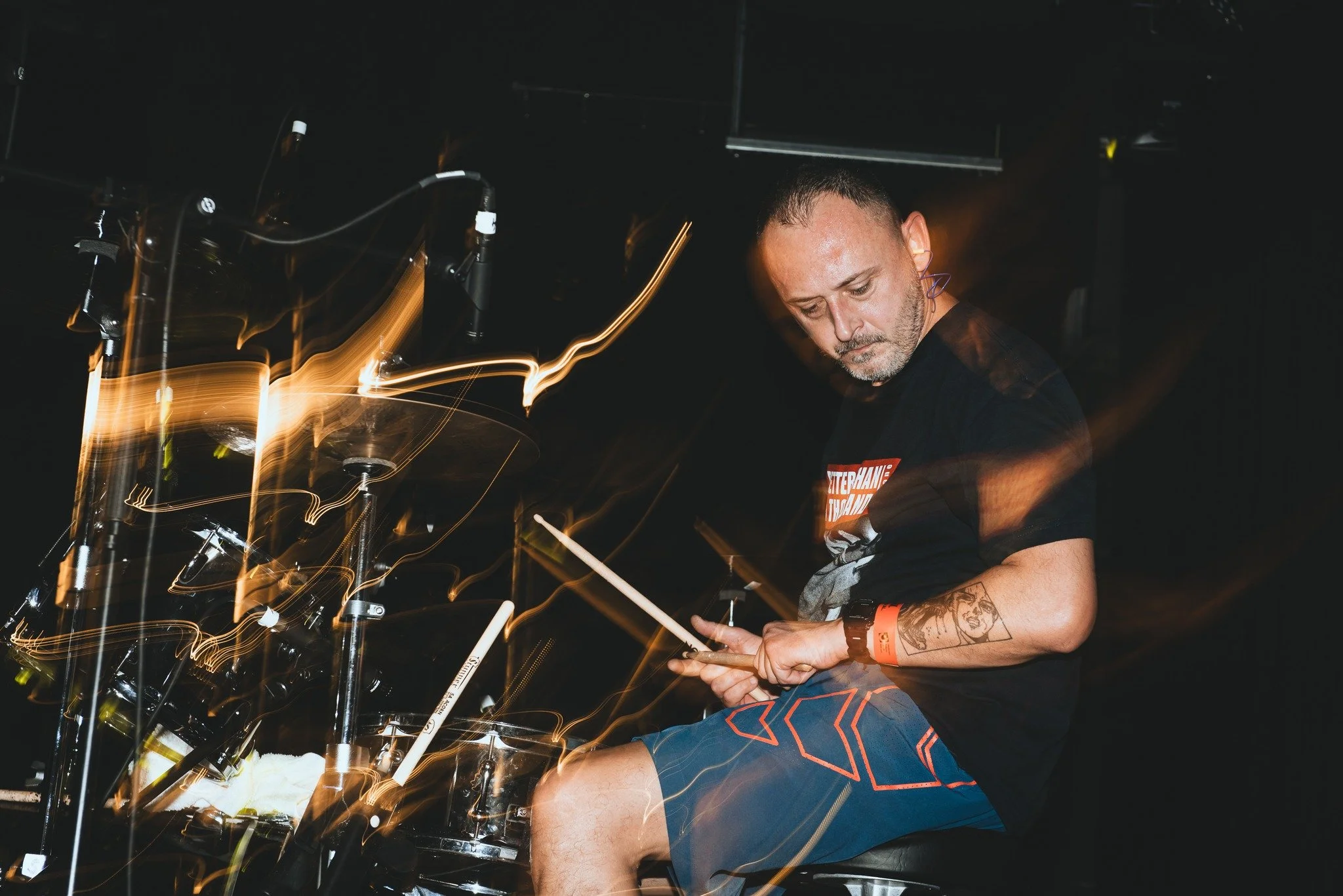 A man with tattoos on his arm, wearing a black T-shirt, is performing as a DJ in a dark environment with light streaks around him, possibly at a concert or nightclub.