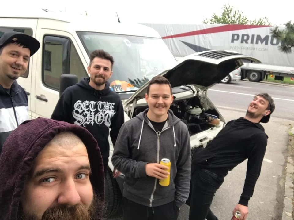 Group of five young men standing on the roadside next to a white van and a truck, with a car engine hood open.