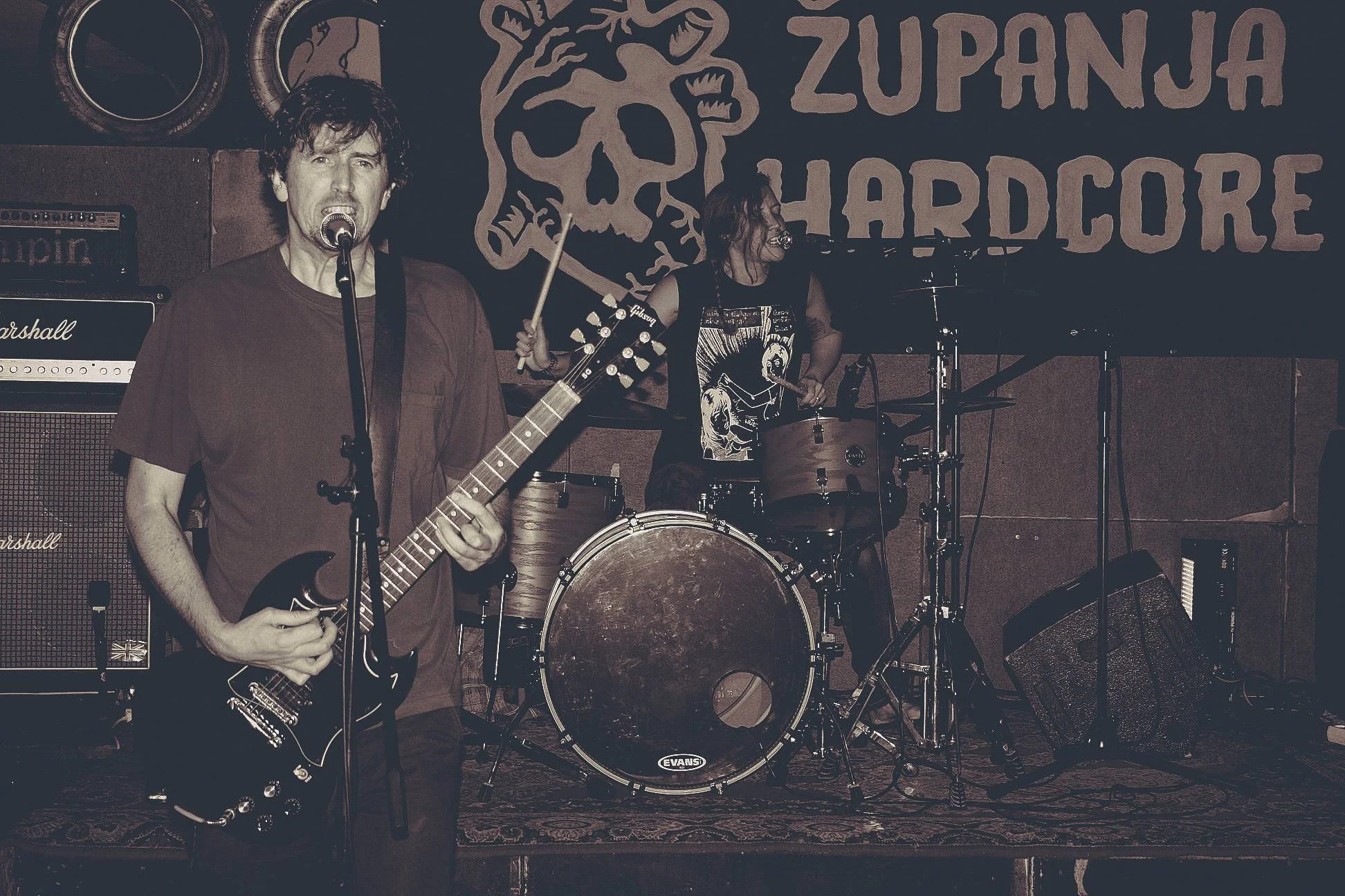 A band performing live on stage, featuring a male guitarist and vocalist with dark curly hair and a female drummer with long dark hair. A backdrop displays a skull and the words "Zupan Hardcore."