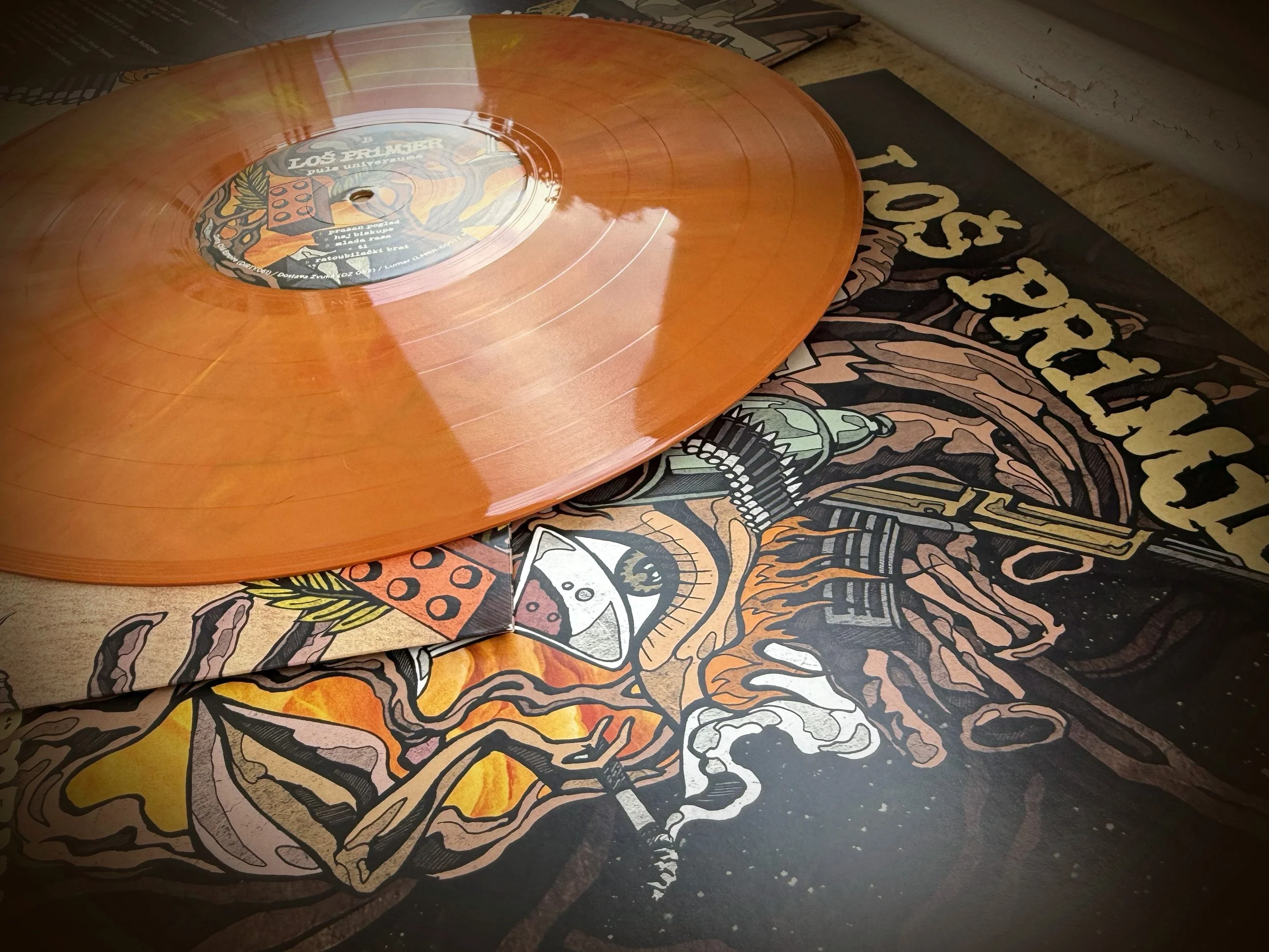 Orange vinyl record on top of a colorful, graphic-illustrated album cover with the word "LOS PRIMJER" visible.