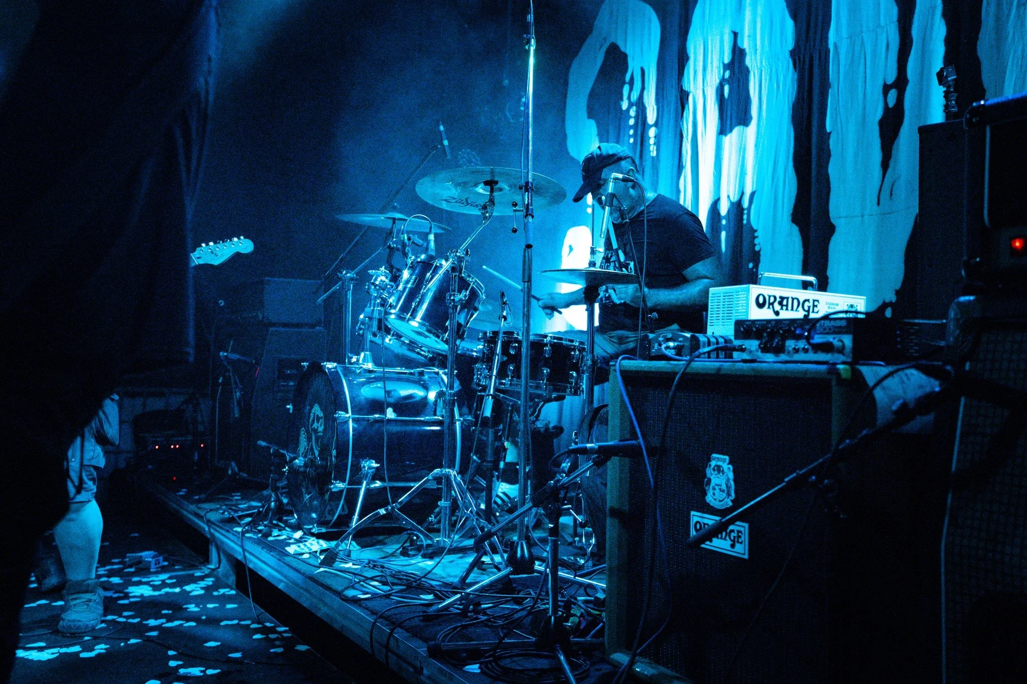 Musician adjusting electronic equipment on stage with drums and guitar, dark blue lighting, and a backdrop with abstract shapes.