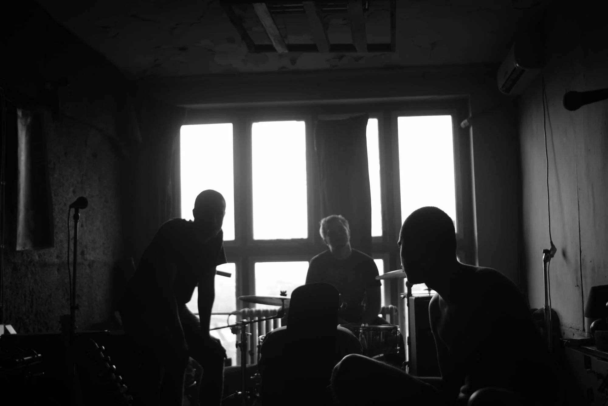 Black and white photo of four people in a room with large window, some sitting at a table, one with a drum kit, diffused sunlight through the window.