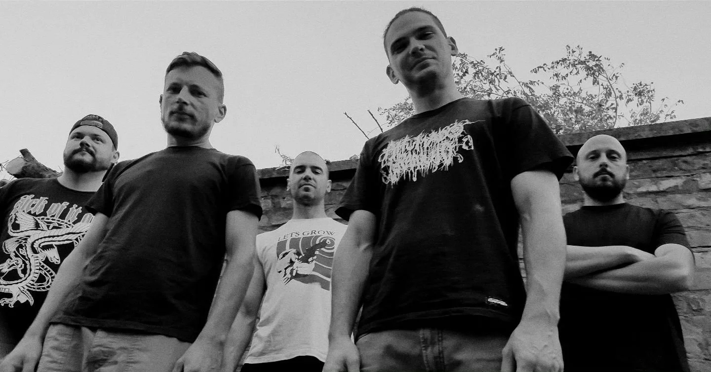 A black-and-white photo of five men standing outdoors in front of a brick wall, looking down at the camera with serious expressions.