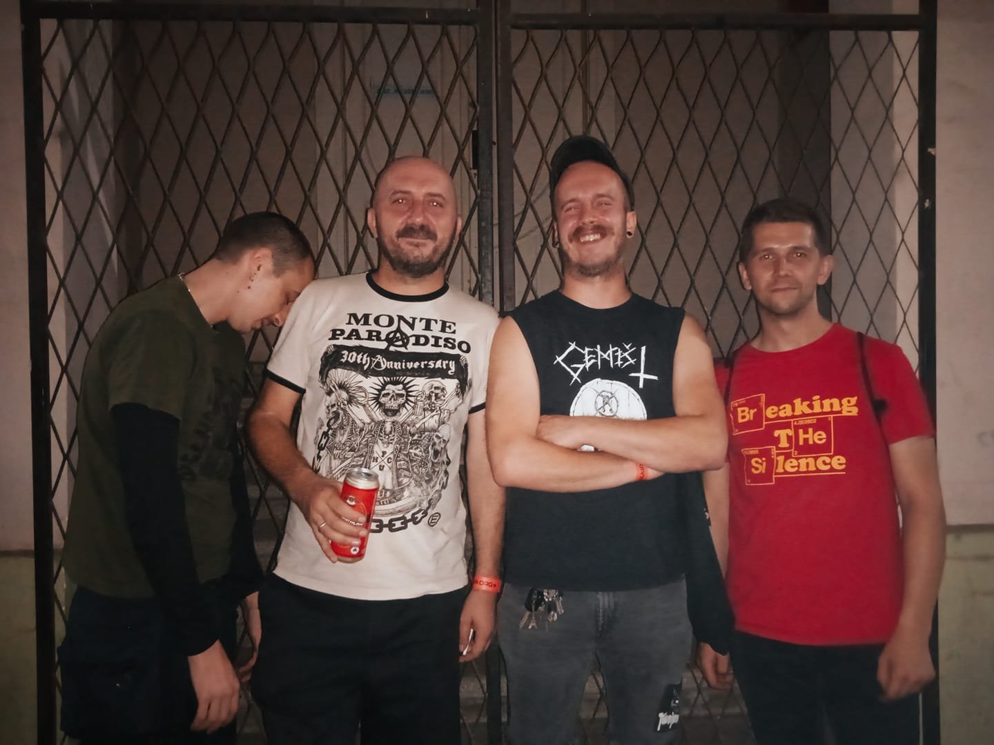 Four men standing in front of a metal gate, with one looking down and the others smiling at the camera. Two are holding drinks, and they are dressed in casual clothing including band and science-themed T-shirts.