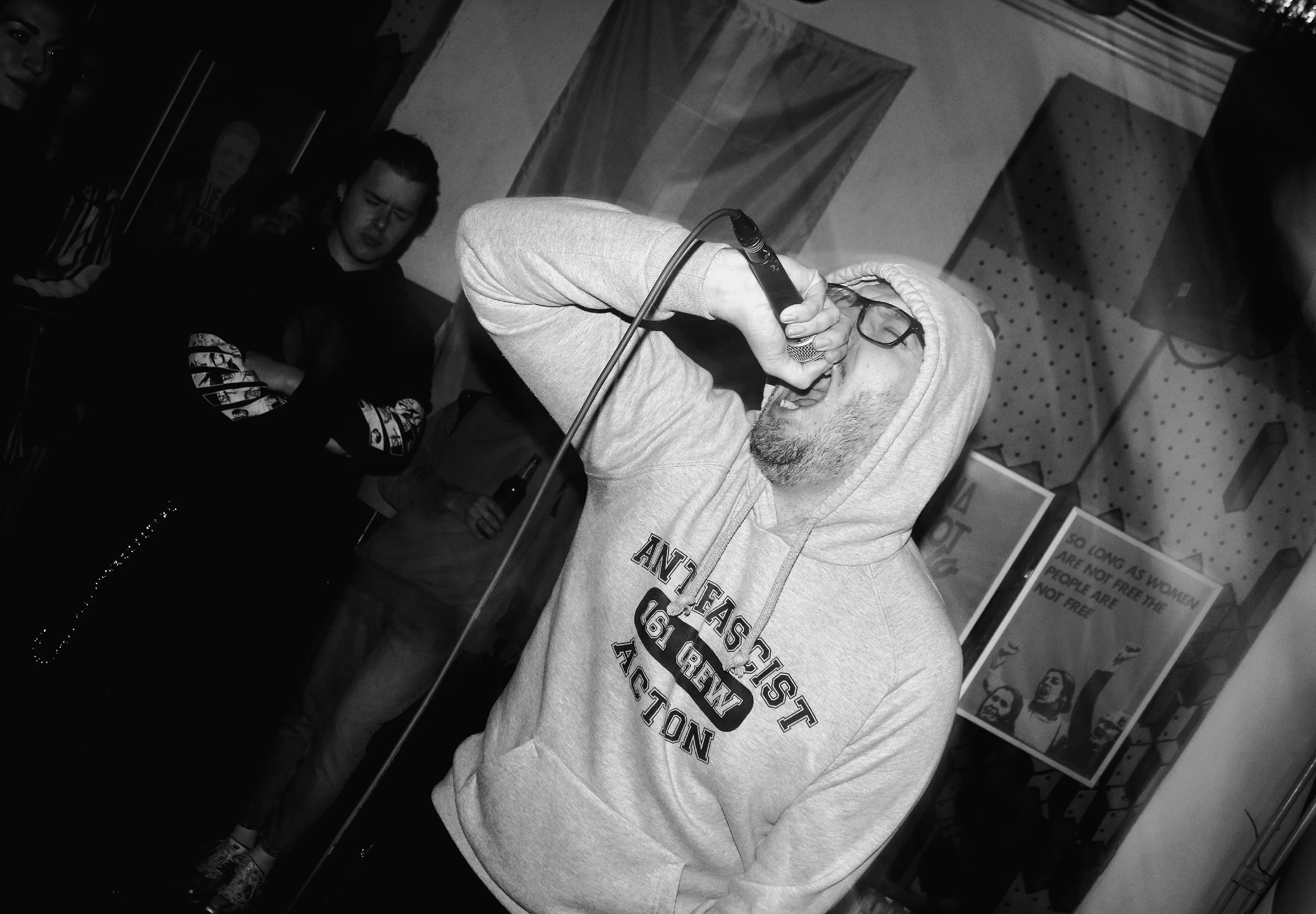 A man with glasses and a hoodie singing into a microphone at an indoor event, with several people sitting behind him and posters on the wall.
