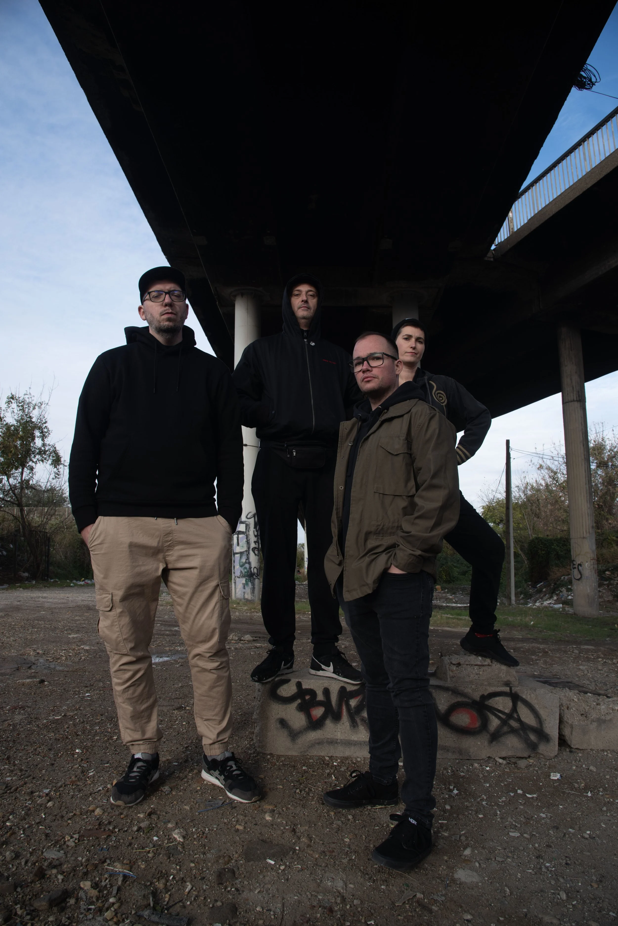 Five young adults standing under a bridge with graffiti on a concrete support, during daytime. The group is casually dressed, with some wearing hoodies and jackets. They pose confidently with serious expressions, with a cityscape and trees in the background.