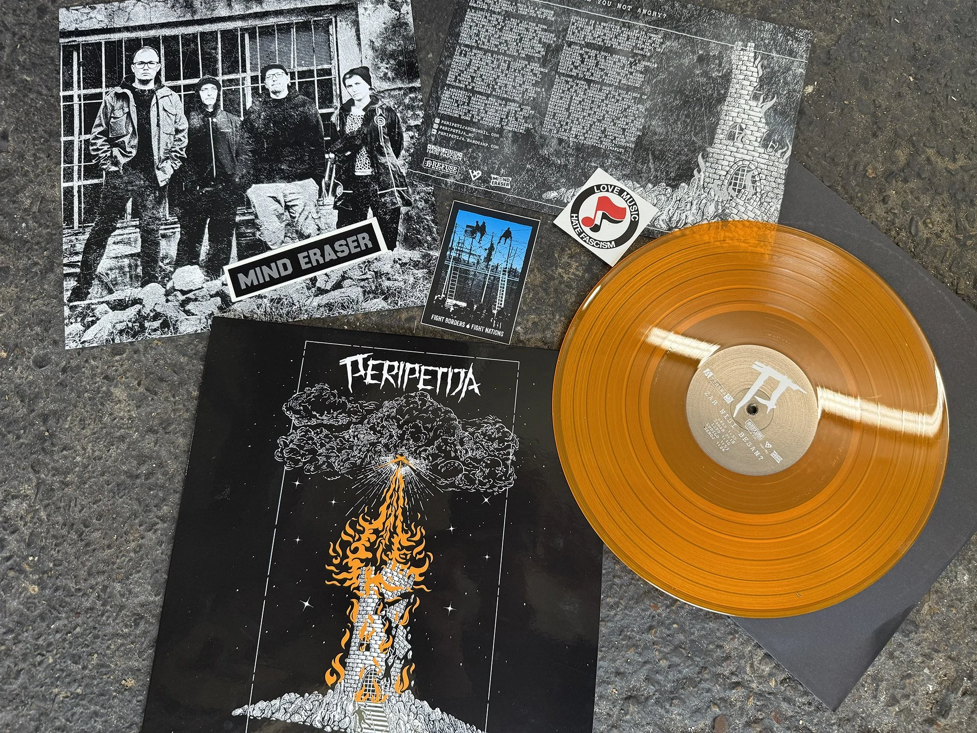An orange vinyl record, black and white band posters, and stickers are laid out on a textured surface. The posters feature band members, text, and artwork, including a volcano eruption illustration.