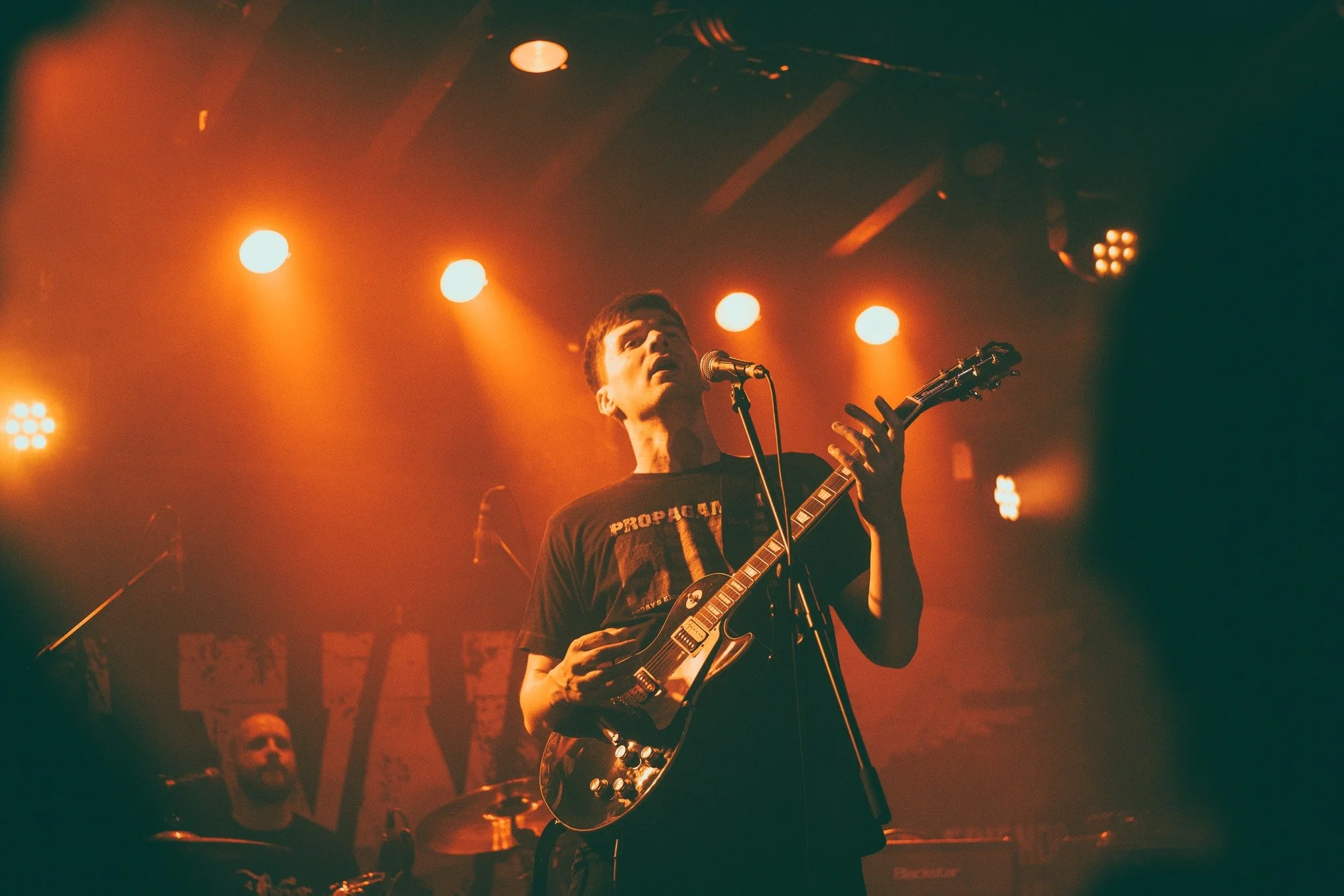 A young male musician singing into a microphone and playing an electric guitar on stage, with orange stage lights illuminating the scene.
