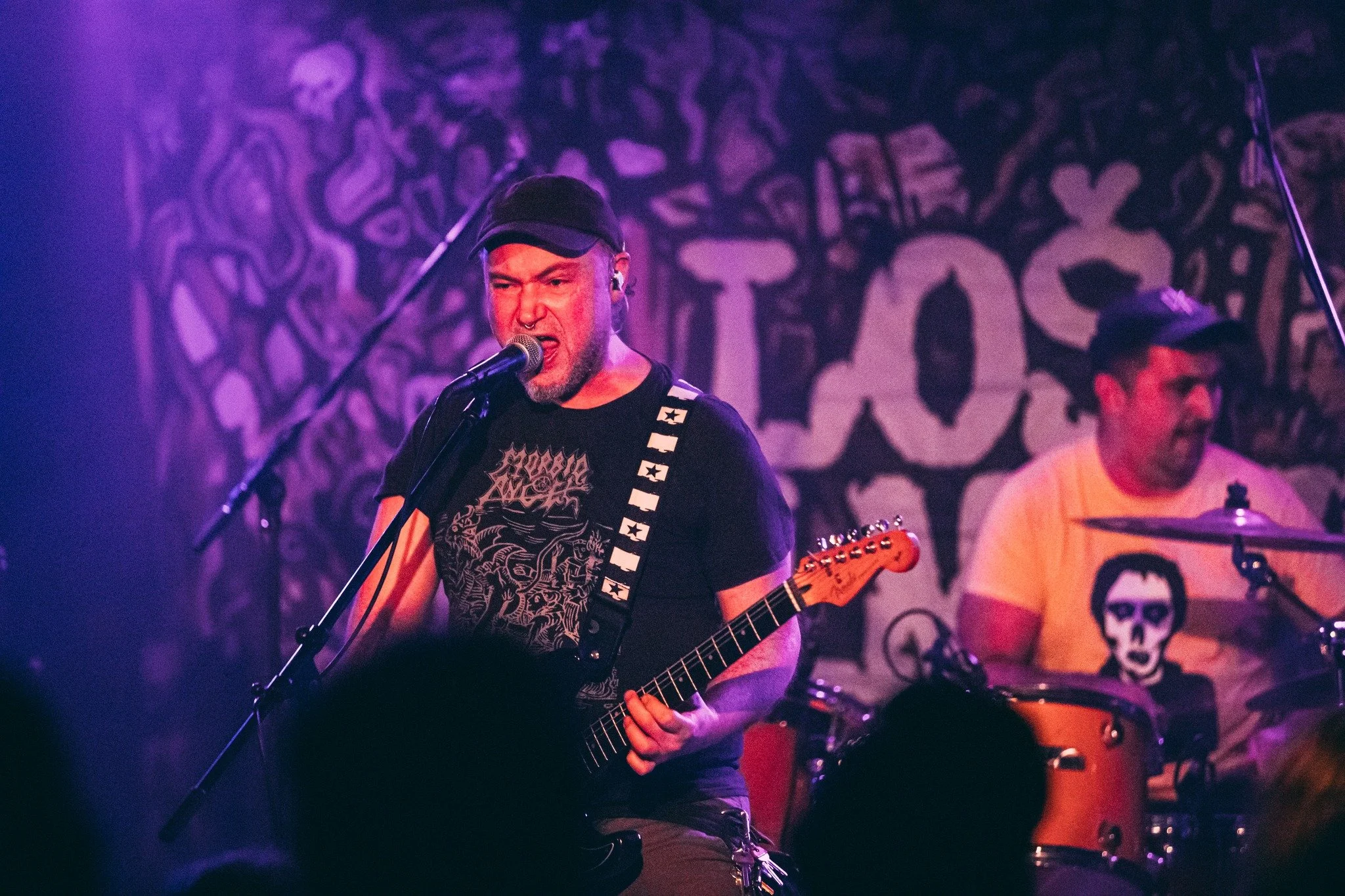 A man with a cap and earrings passionately singing into a microphone and playing an electric guitar during a live music performance, with a second musician visible in the background behind a drum set.