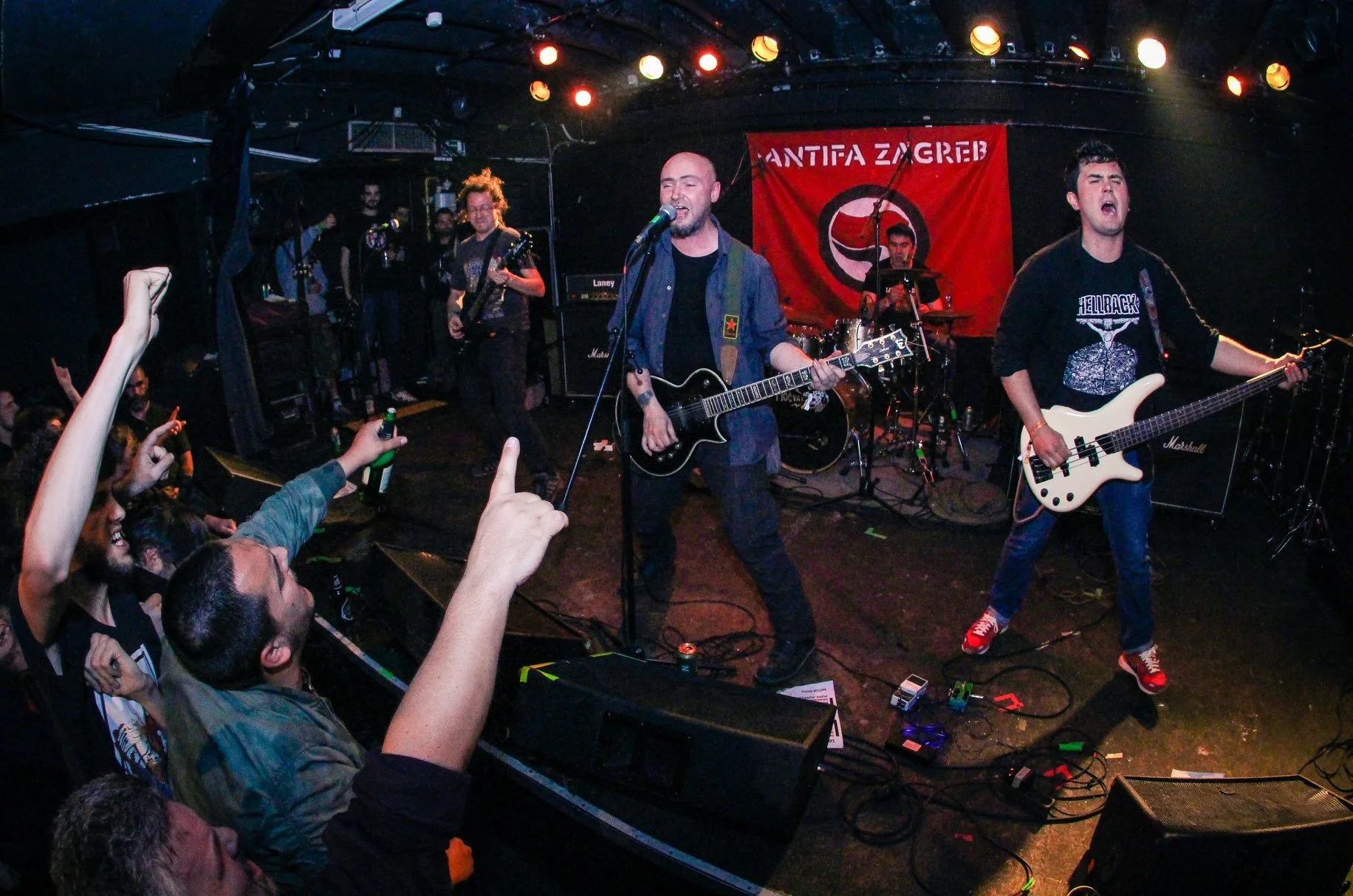 A live band performs on stage at a concert venue with an audience nearby. The band consists of a singer with a guitar, a bassist, a guitarist, and a drummer. A red banner with black and white symbols hangs behind them.