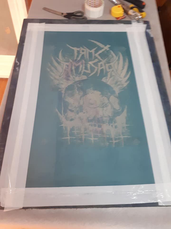 A screen printing frame with a stencil of a skull wearing a hat, with stylized text above and a decorative background. The frame is on a table with tools including scissors, tape, and a utility knife.