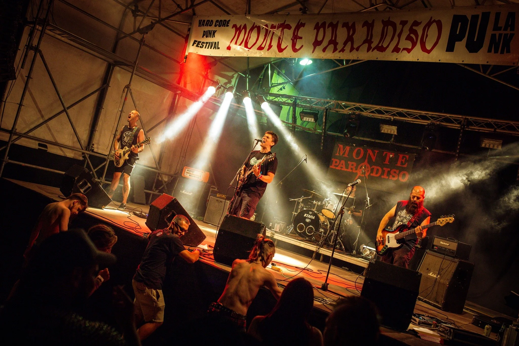 A live band performing on stage at a music festival, with musicians playing guitars and drums under stage lighting and a banner that reads 'Monte Paradiso' and 'Hard Core Punk Festival'.