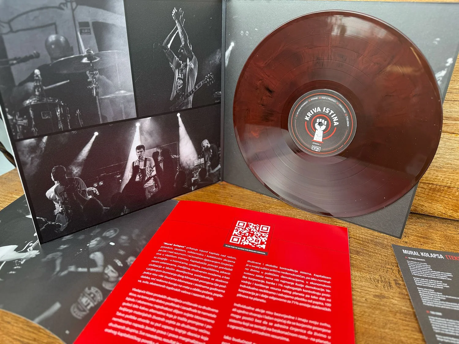 A vinyl record with an orange marbled appearance in a black sleeve, accompanied by black and white photographs of a band performing live and printed informational material on a wooden surface.