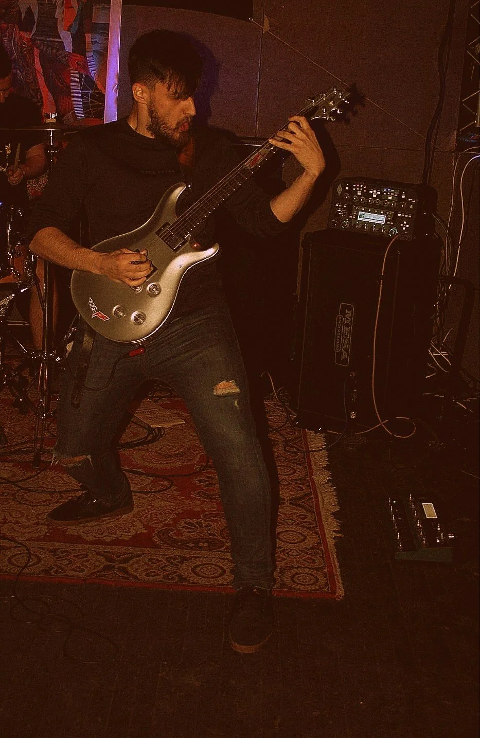 A man playing an electric guitar on stage during a live music performance, with music equipment and a drum set visible in the background, and a patterned rug on the floor.