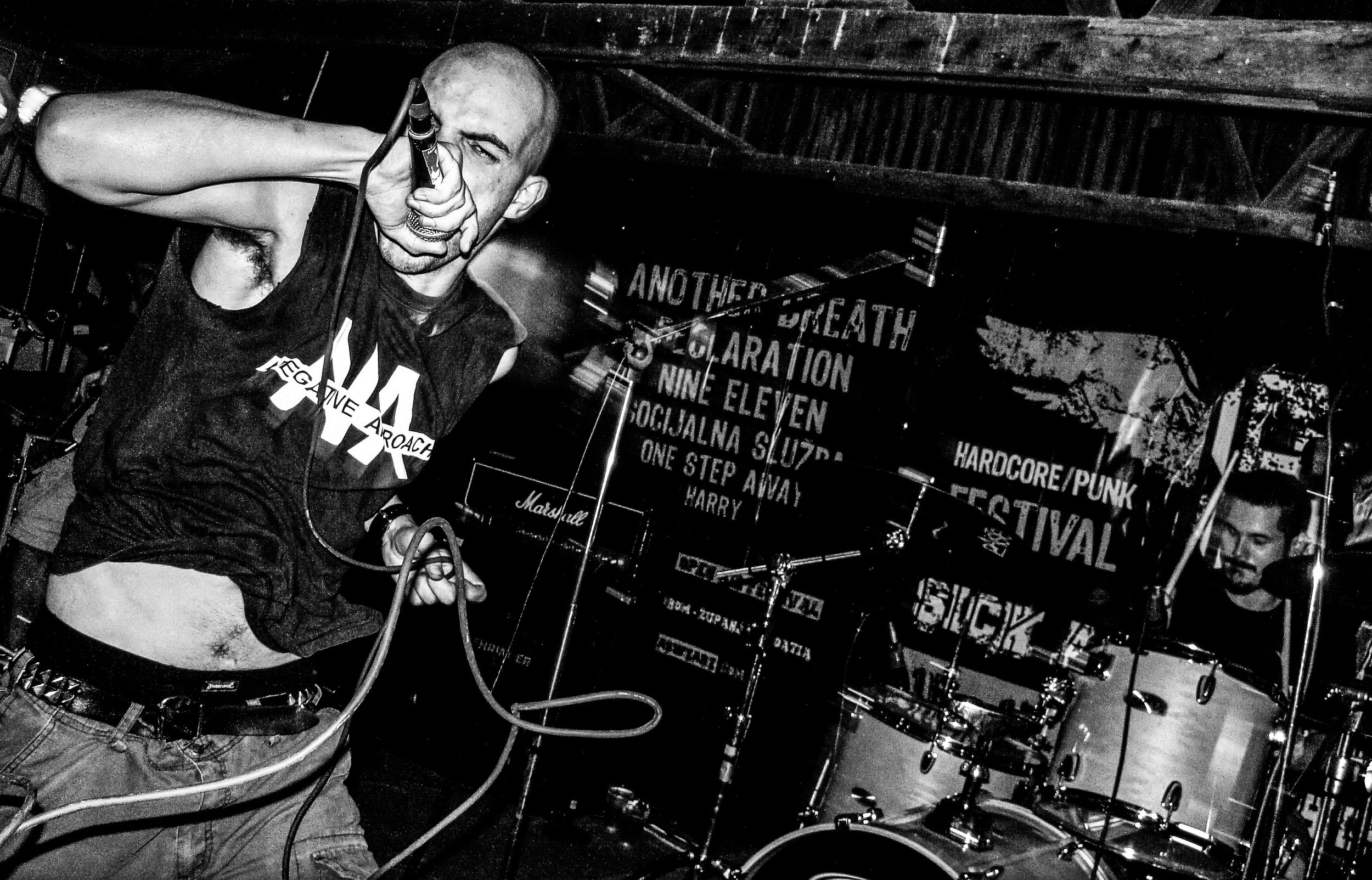 A bald man with tattoos on his arm is singing into a microphone at a live music performance. He is wearing a sleeveless shirt with text on it. There are musical equipment and amplifiers around him, and a banner with band or event names in the backgro