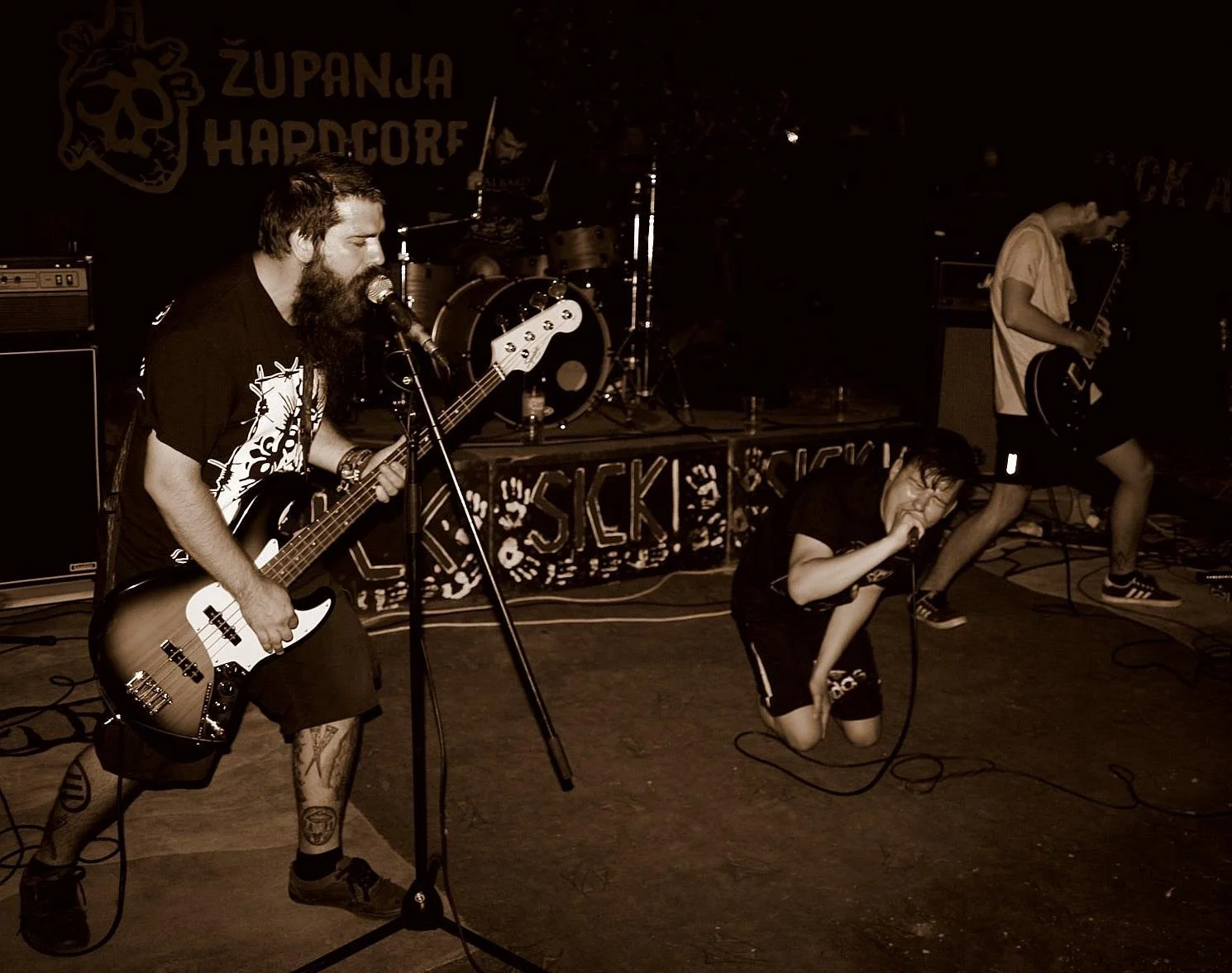 A band performing on stage with three members. One with a beard singing into a microphone and playing an electric bass guitar, one in dark clothing kneeling and singing into a microphone, and one with a guitar in a white shirt and shorts standing in 
