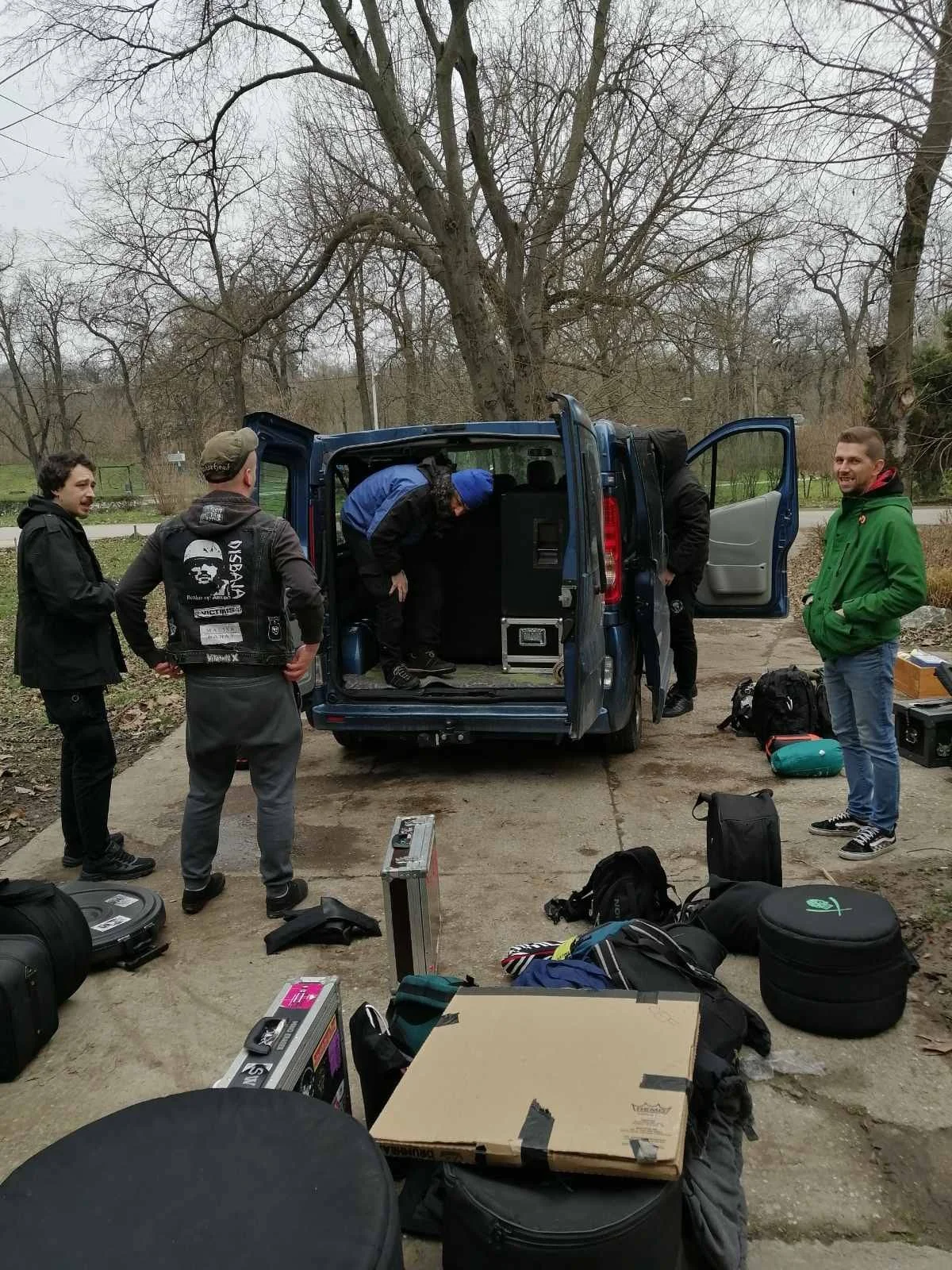 A group of five men preparing for an outdoor activity, with gear and supplies laid out around them near a blue van in a park-like setting with leafless trees.