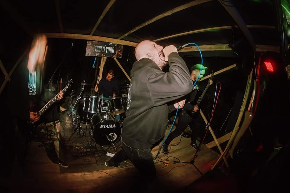 A band performs live on stage at a small venue. The lead singer is bald, singing into a microphone, and wearing a dark hoodie. To the left, a guitarist in black attire is playing, and in the background, a drummer is playing the drums. The stage has w