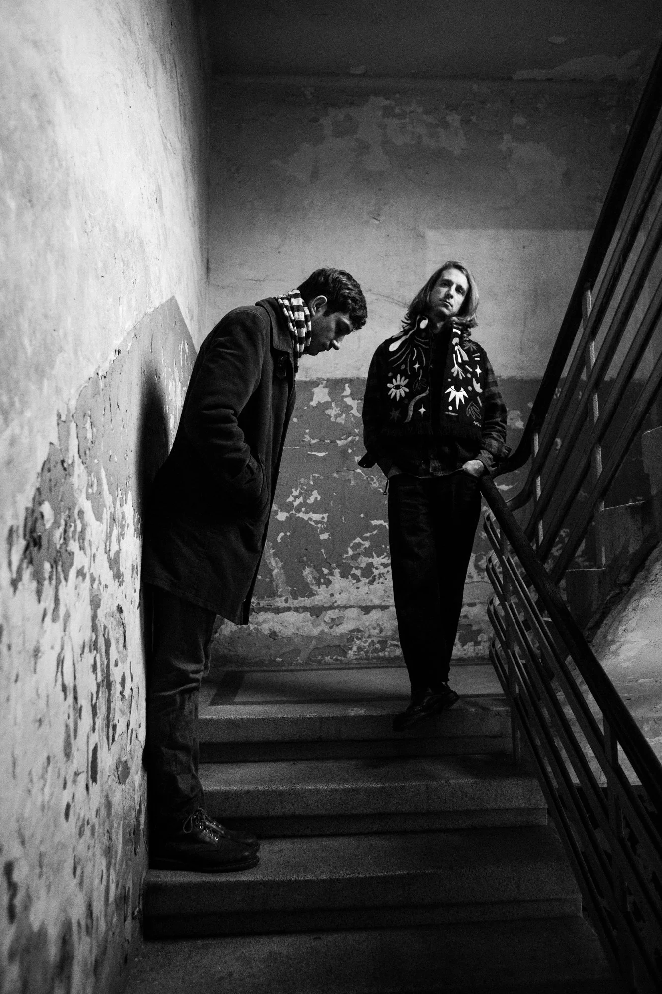 Two people standing on an indoor staircase with worn walls. One person is leaning against the wall with head bowed and hands in pockets, the other is standing with hands in pockets, looking slightly upward.