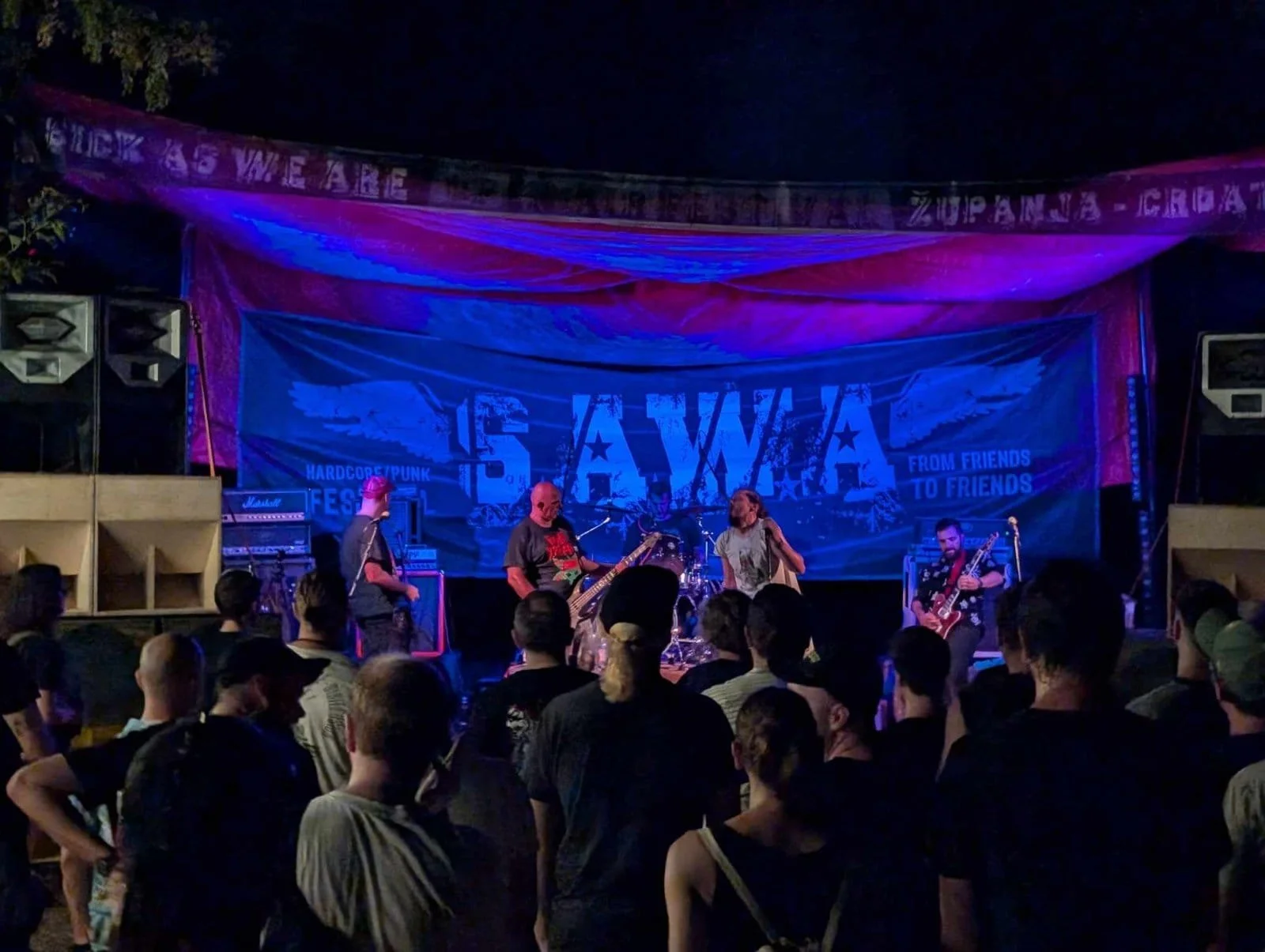 Band performing on an outdoor stage at night with a crowd watching, a banner displaying 'SAWA' and musical equipment visible.