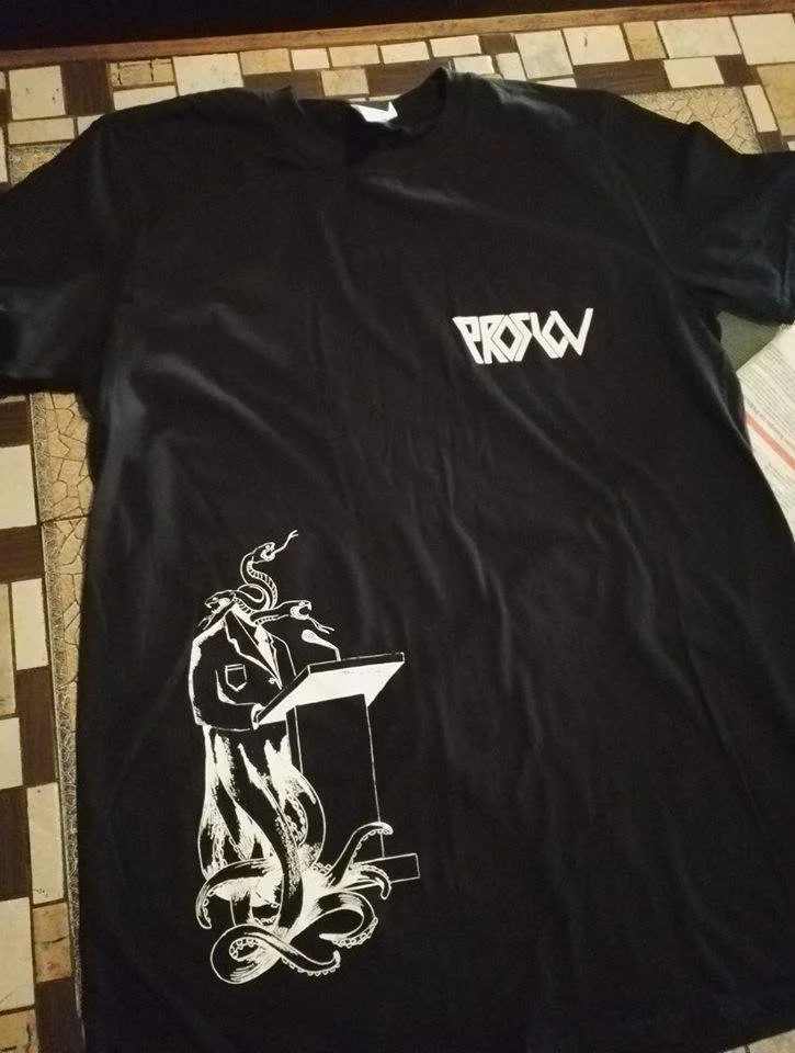 Black T-shirt with the logo 'PROSLOV' on the upper right chest area and a white graphic of an anthropomorphic snake creature wearing a jacket, standing at a podium with a microphone.