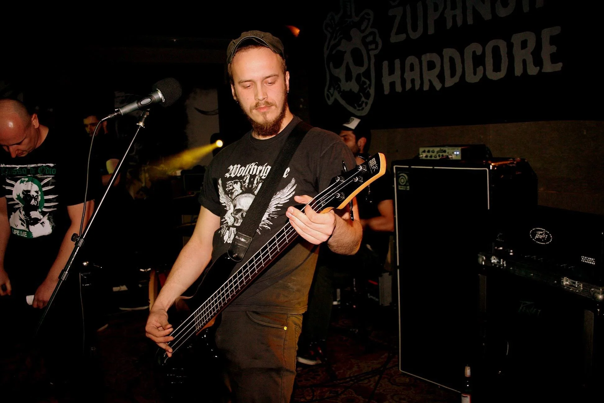 A young man with a beard playing a bass guitar on stage during a live music performance, with sound equipment and a band in the background