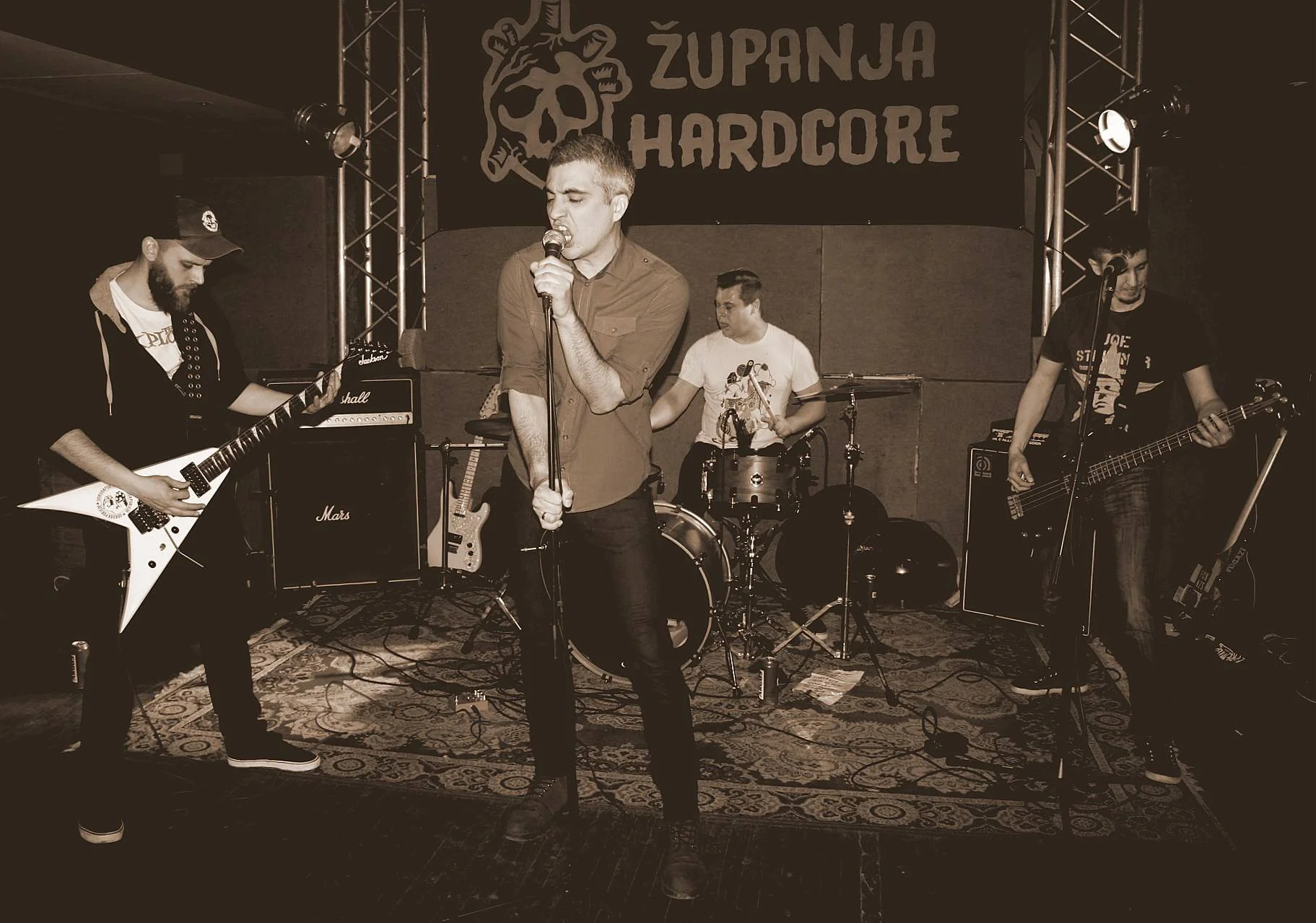 A band performing on a concert stage with a banner that says 'Županja Hardcore'. The band includes a lead singer, a guitarist playing a white Flying V guitar, a drummer, and a bassist. The scene is in black and white, with musical amplifiers and equi