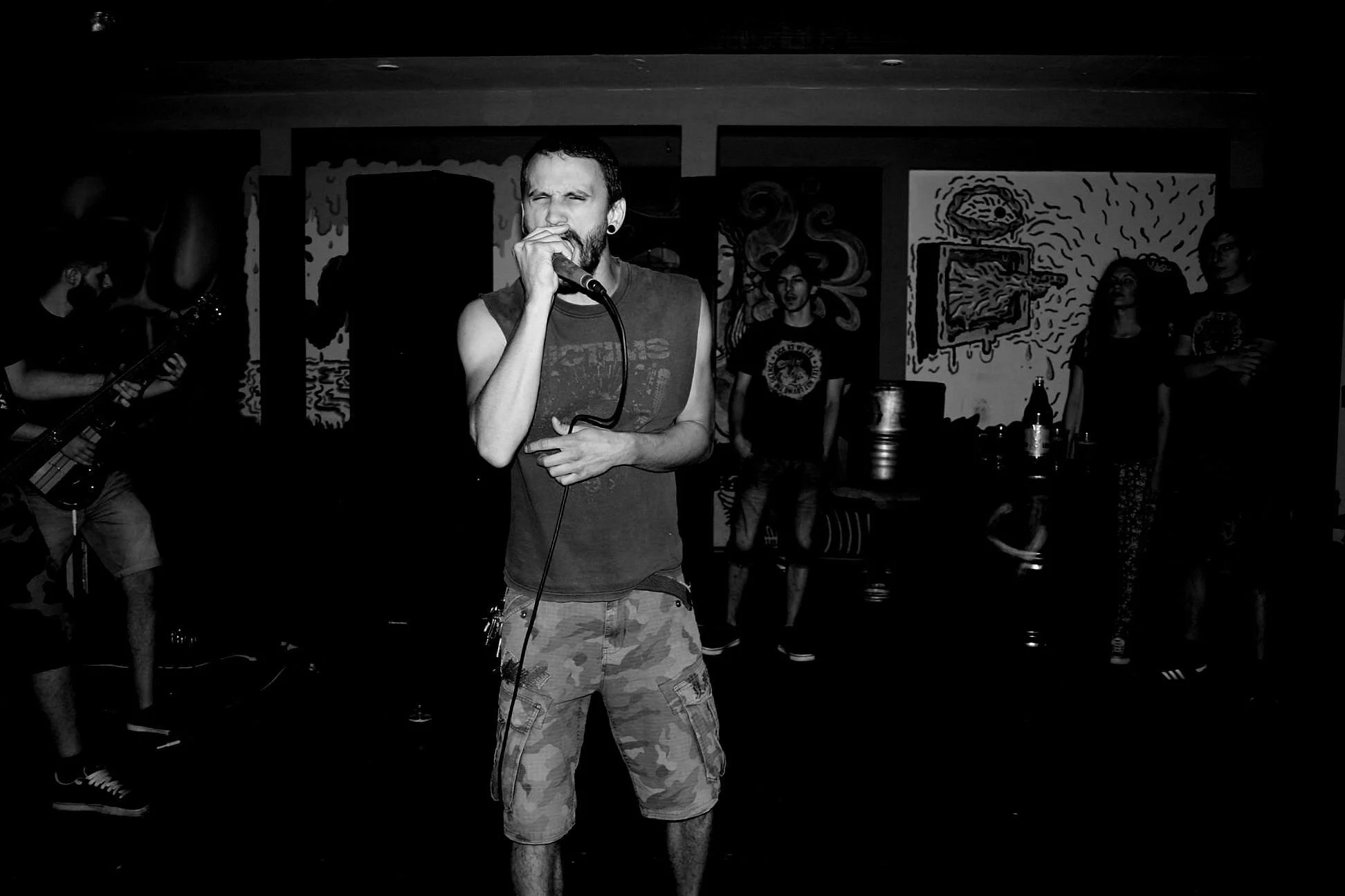 A black and white photo of a man with a beard and short hair singing into a microphone at a music event, wearing a sleeveless t-shirt and camouflage shorts, with a band or audience in the background.