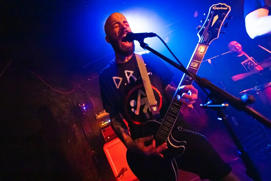 A male musician passionately performing live on stage with an electric guitar, singing into a microphone, in a dimly lit venue with colorful stage lights.