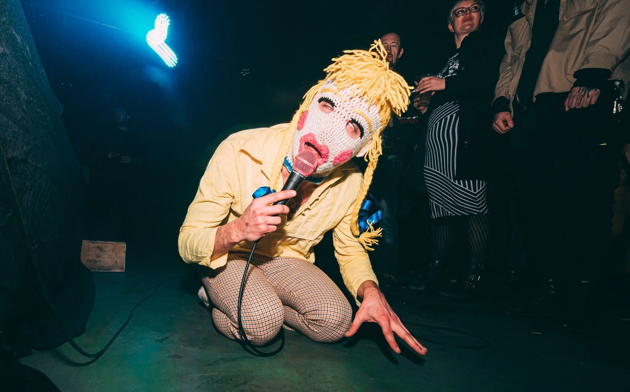 Person with a knitted clown mask, yellow yarn hair, and fishnet stockings performing with a microphone at a dark event, surrounded by a small audience.