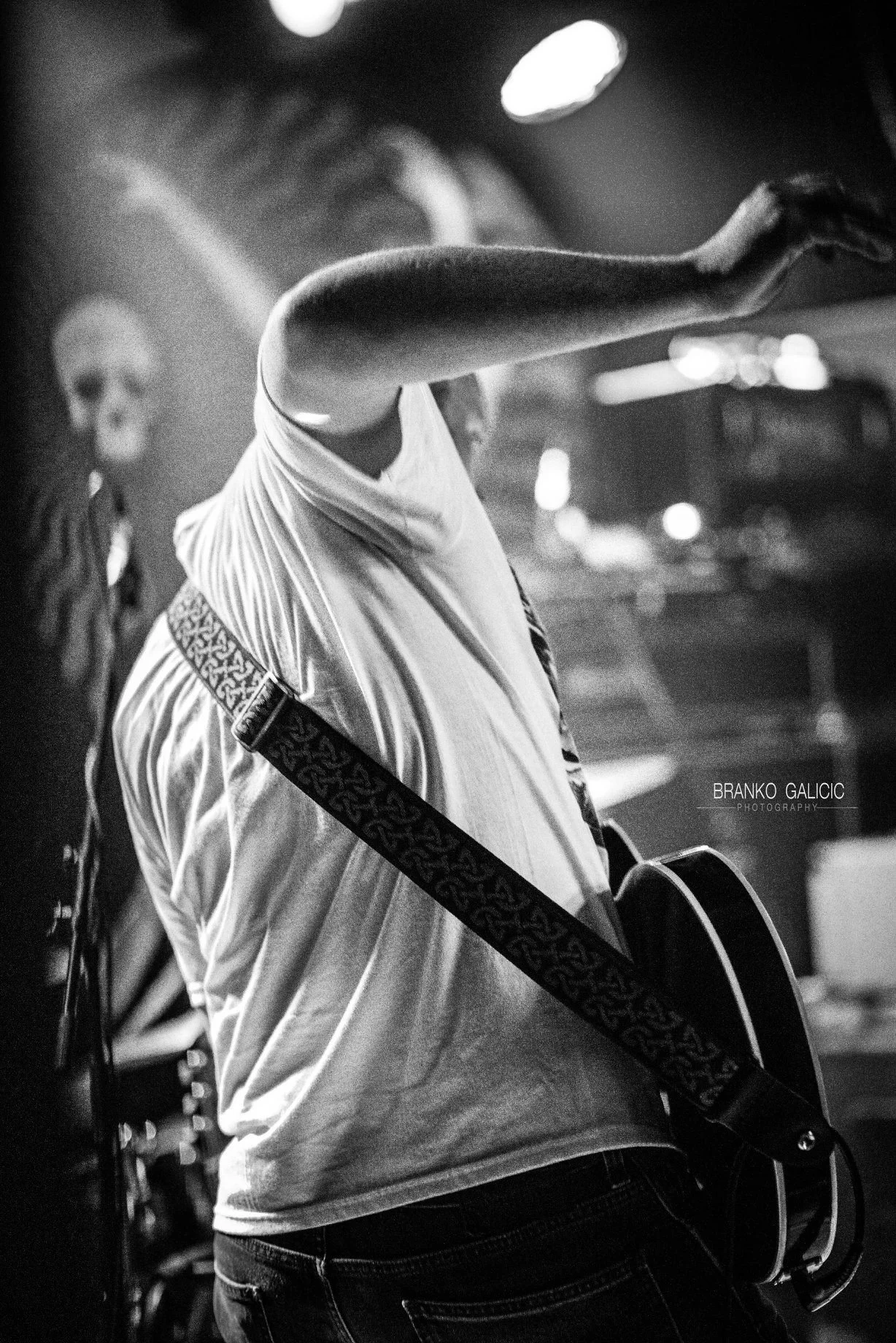 A person playing a guitar on stage at night, wearing a light-colored t-shirt, with a skateboard strap hanging from their shoulder, and another person with long hair is partially visible in the background.