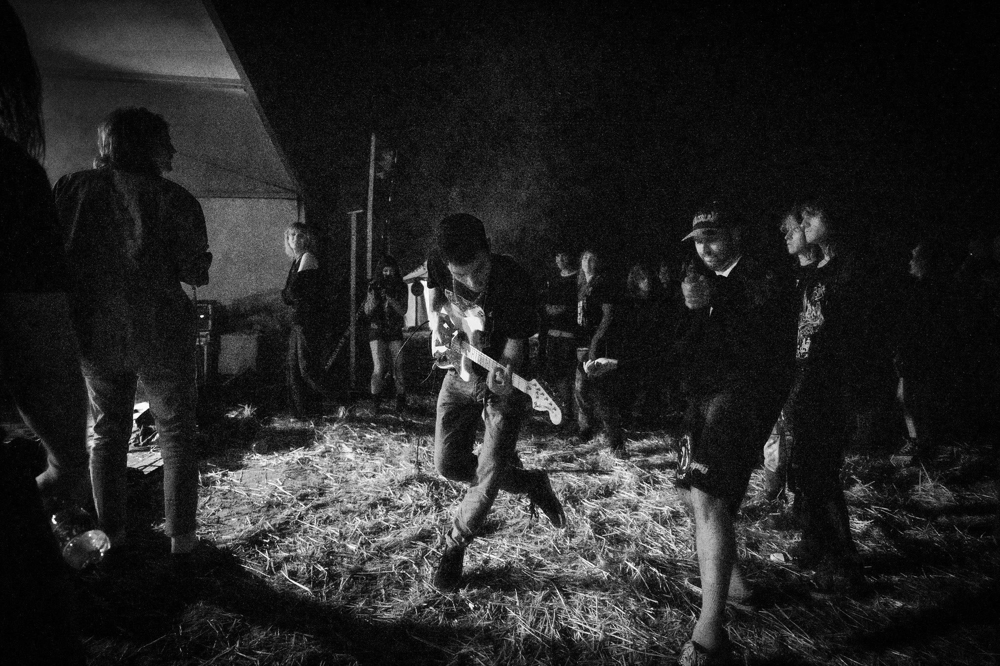 A group of people gathered outdoors at night, some playing musical instruments and others standing or sitting around. The scene appears to be lit by a nearby fire or light source, with the ground covered in straw or dry grass.