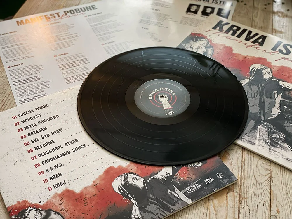 A black vinyl record on top of a printed album cover and booklet, with a list of song titles in Croatian, all on a wooden surface.