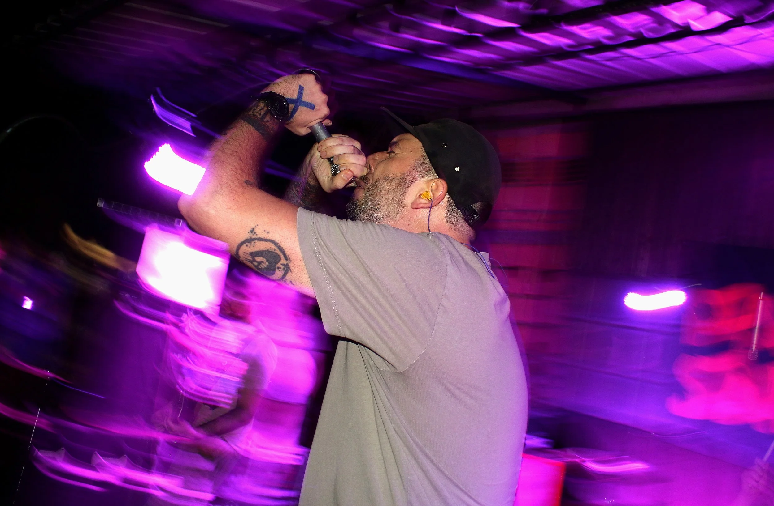 A man singing into a microphone in a dimly lit space with purple and pink lights, wearing a black cap and sporting tattoos on his arms.