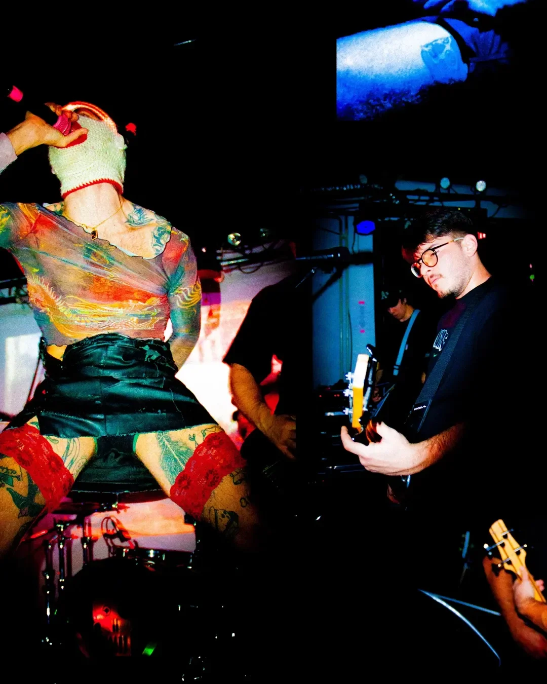 A woman singing into a microphone on stage, wearing a colorful shirt, black shorts, and a knit hat, with tattoos visible on her arms and legs. Two men are playing guitars nearby in a dimly lit environment with stage lights.