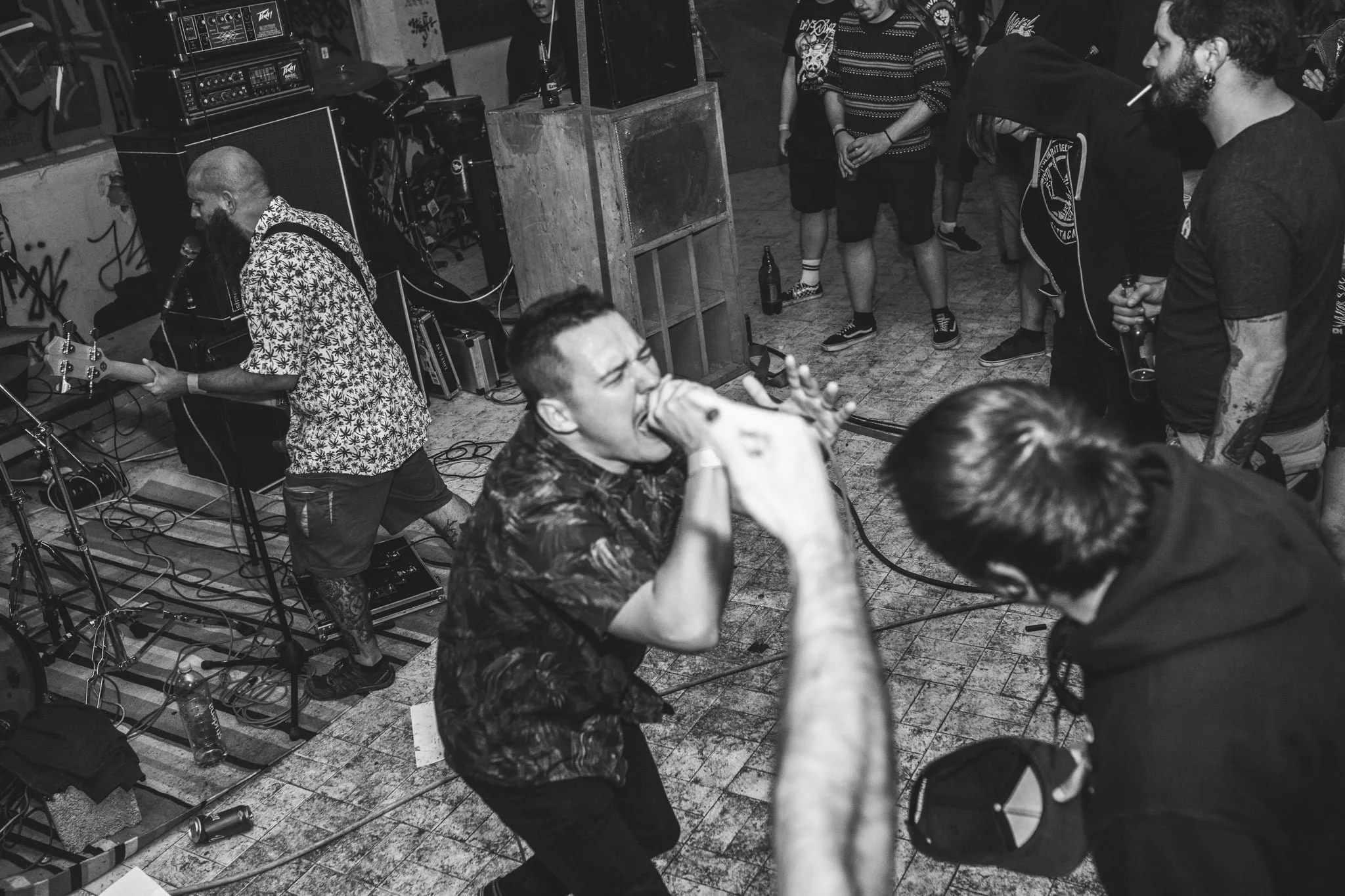Black and white photo of a music rehearsal or concert scene with musicians and audience members, including a man playing guitar, and others singing or watching in a crowded indoor venue.