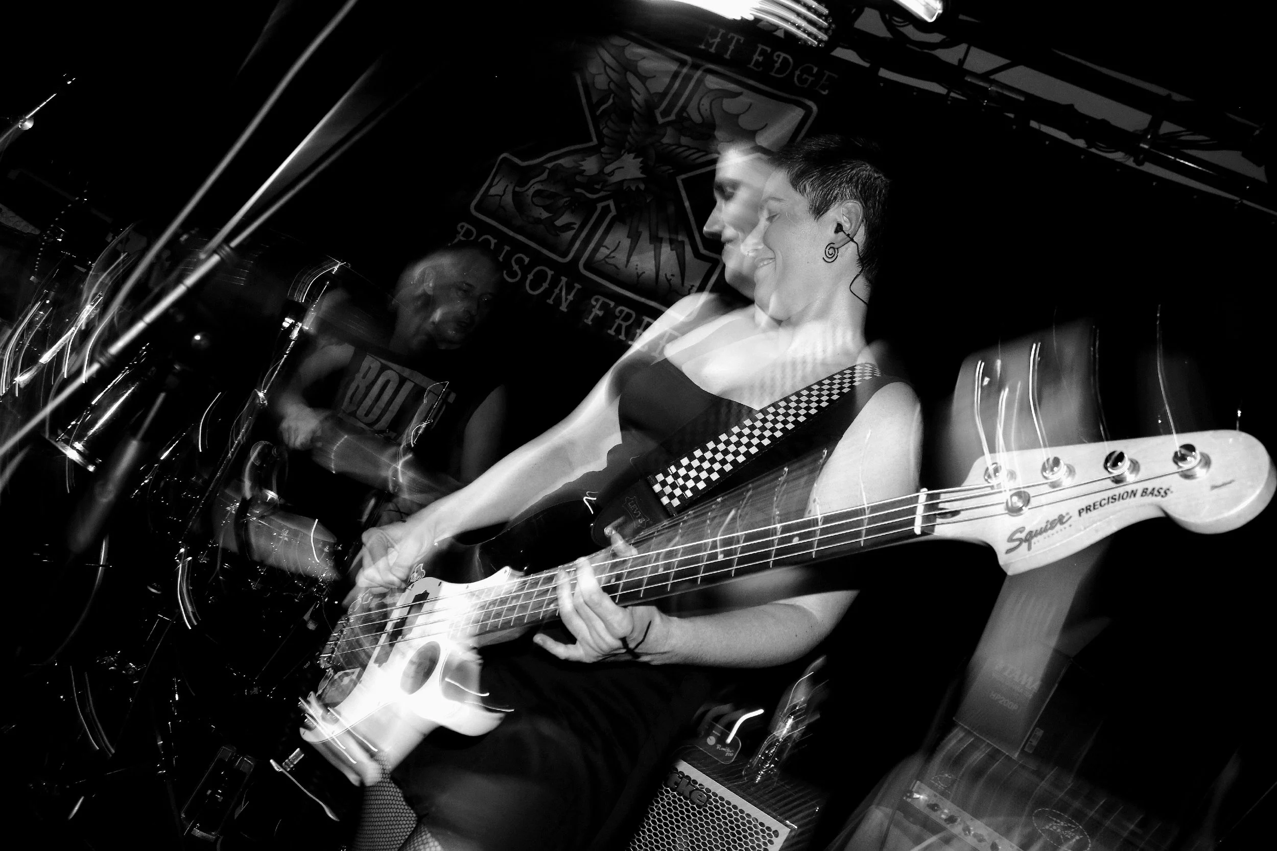 A woman with short dark hair wearing a sleeveless top with checkered straps playing an electric bass guitar with a sticker on the headstock that reads 'Squier Precision Bass'. She is smiling with eyes closed, wearing an in-ear monitor. Behind her, a 